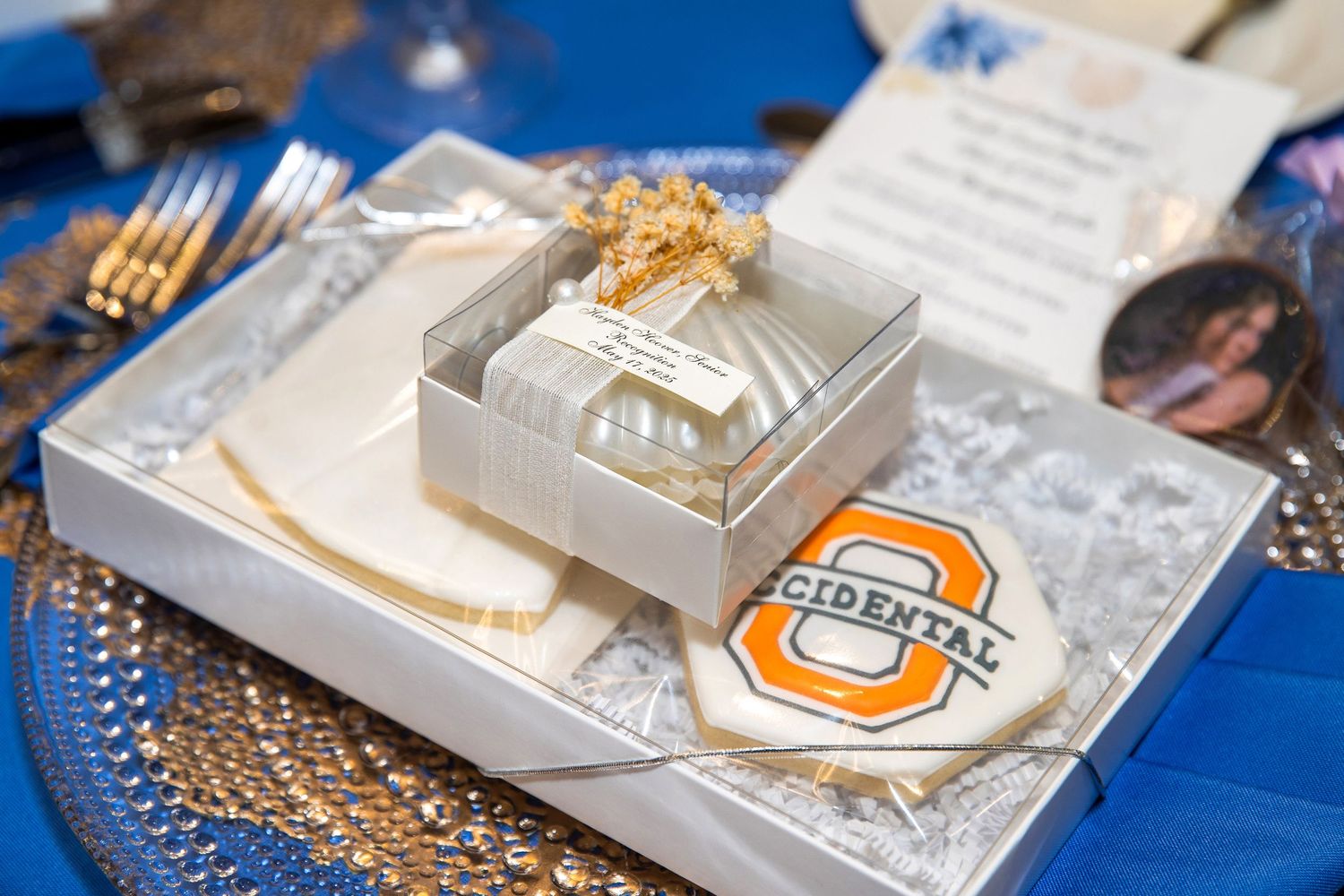 Occidental College branded wedding favor box with gold bow on blue tablecloth with gold charger plate.