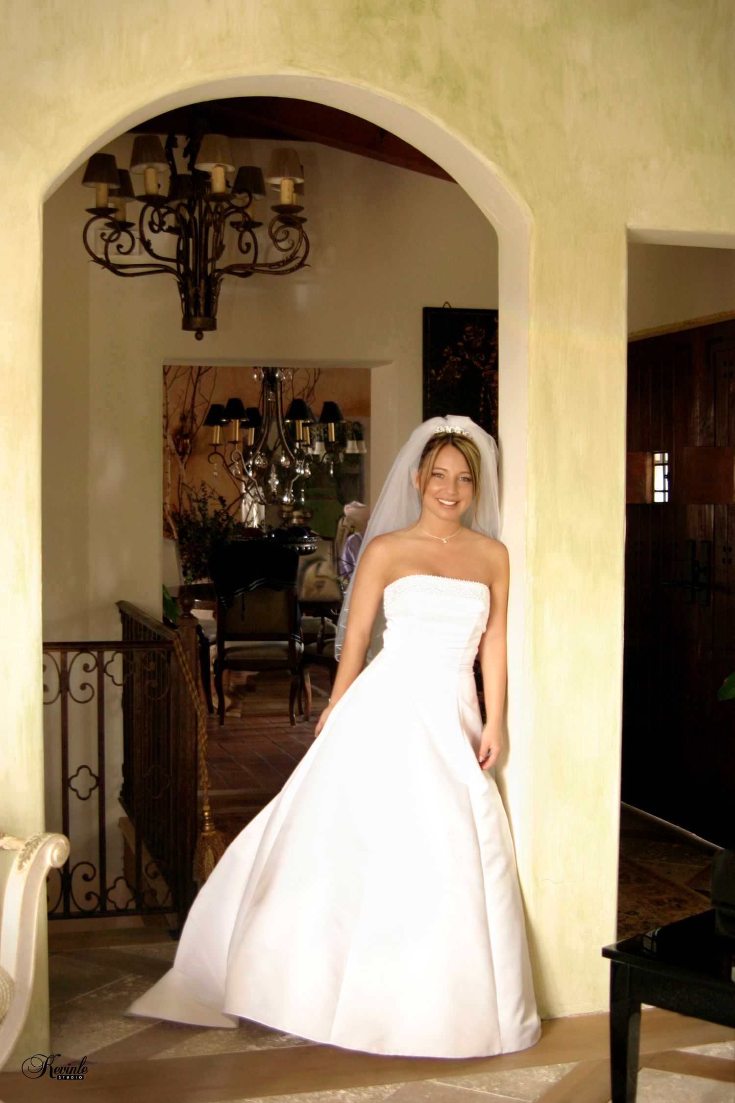 Smiling bride in strapless white gown and veil stands in arched doorway of elegant venue with chandelier.