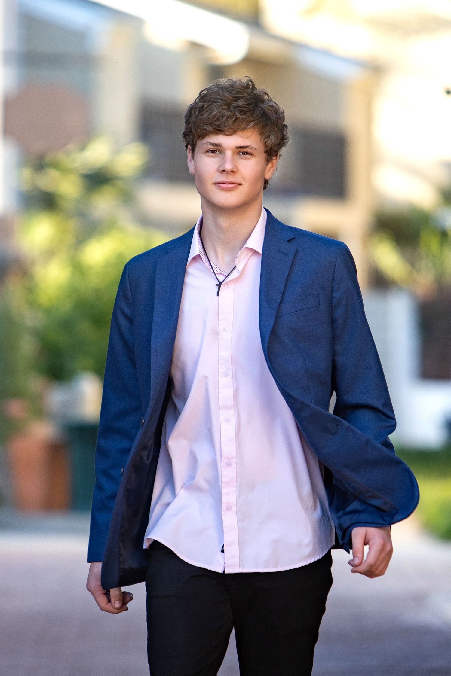 Young man in blue blazer and pink shirt walks outdoors, smiling confidently in a smart casual outfit.