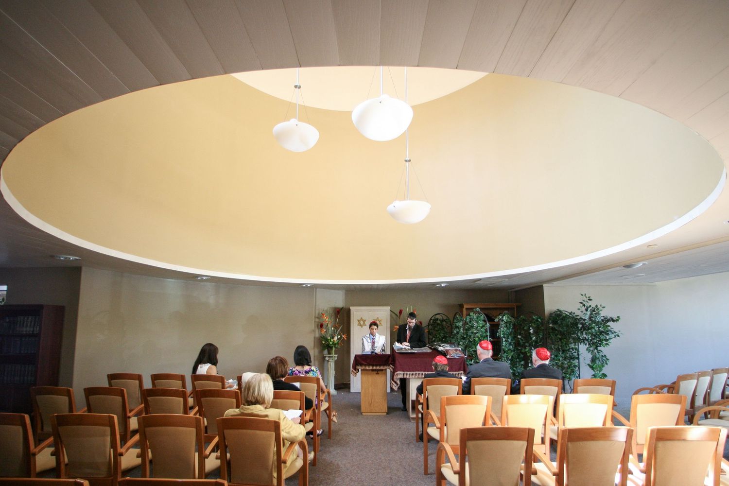 Elegant chapel interior with oval ceiling, warm lighting, wooden chairs, and lush green plants for a serene ceremony space.
