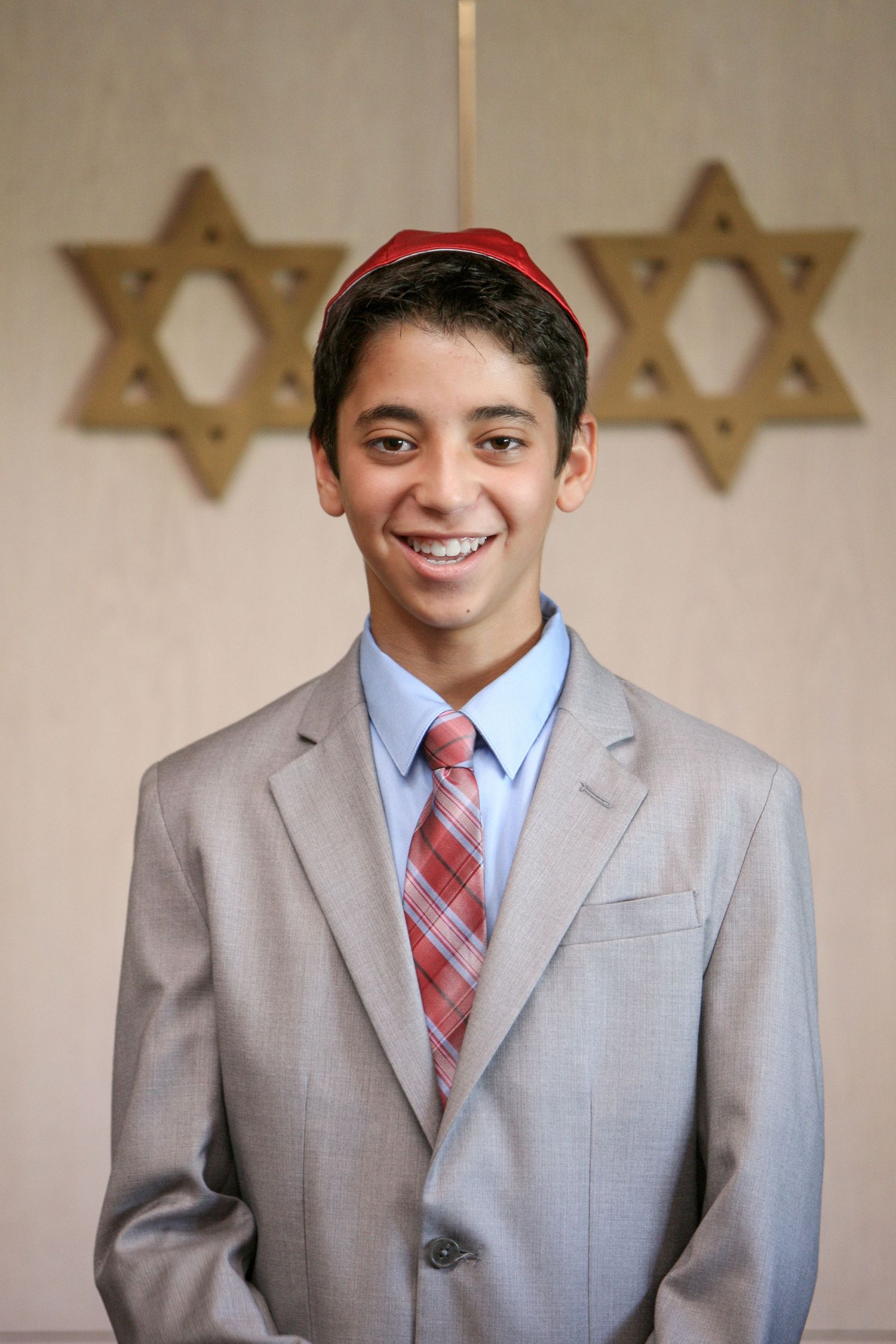 Smiling teenage boy in gray suit and red kippah at Bar Mitzvah ceremony with Star of David backdrop.