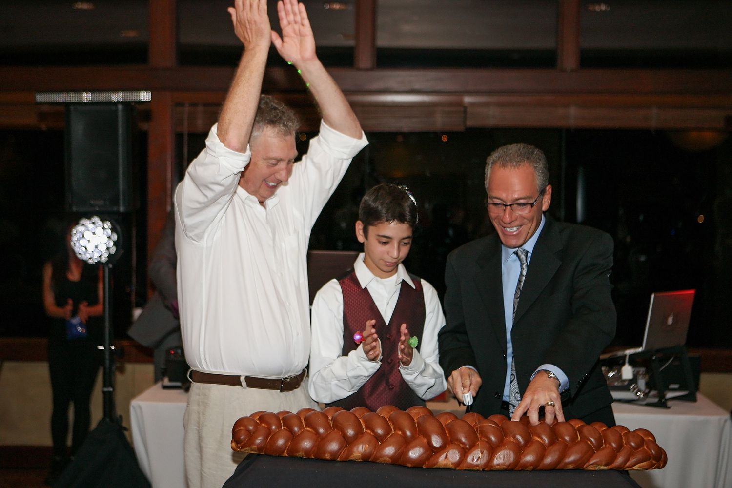 Two men and a boy celebrate at a bar mitzvah party with challah bread on a decorated table.