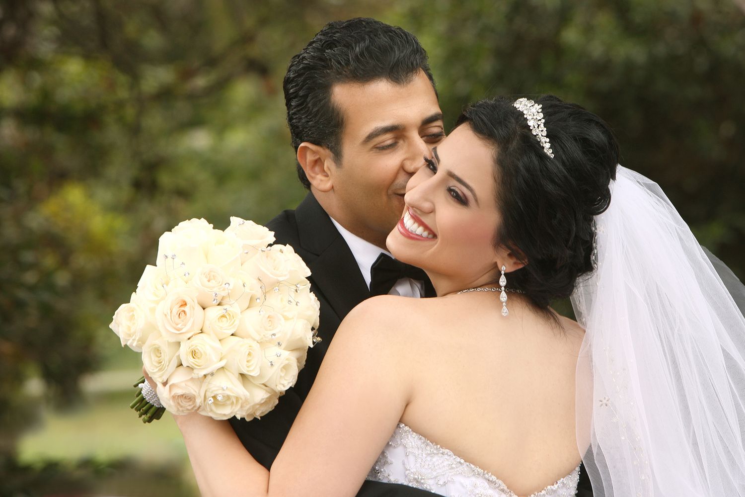 Happy bride holding white rose bouquet while groom lovingly embraces her from behind outdoors.