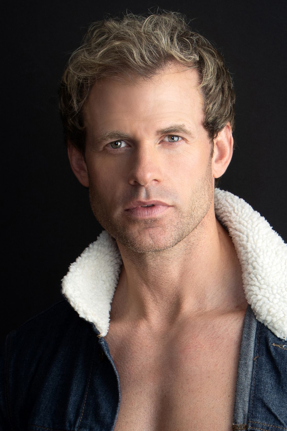 Color portrait of a muscular blond man with blue eyes wearing an open sherpa-lined denim jacket.