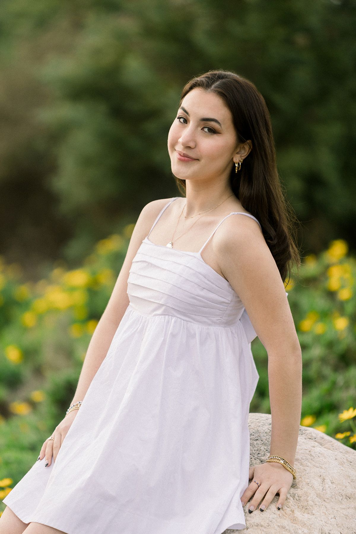 Young woman in white sundress leans on rock surrounded by yellow flowers and green foliage.