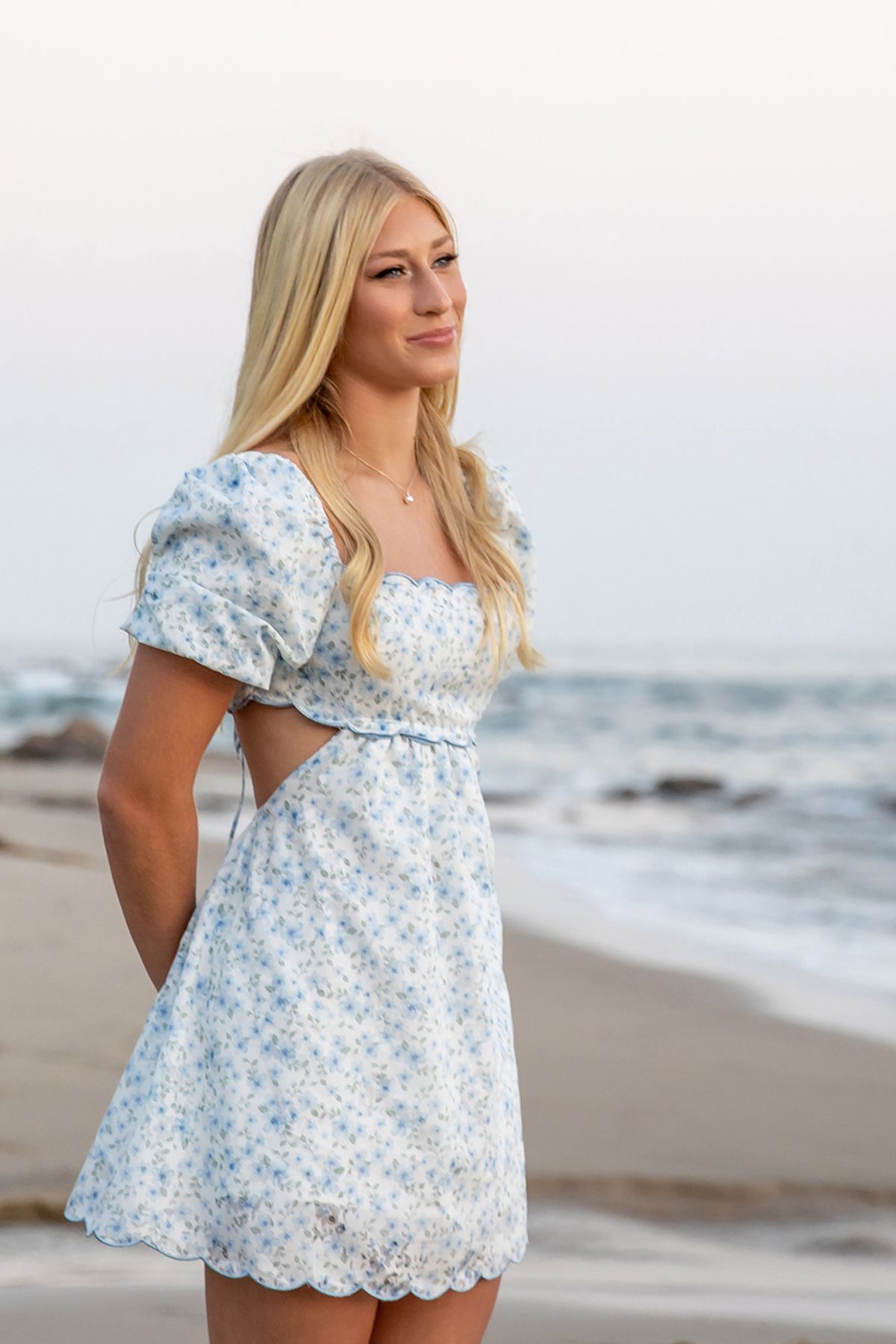 Blonde woman in white blue floral mini dress posing on beach at dusk with ocean waves behind.
