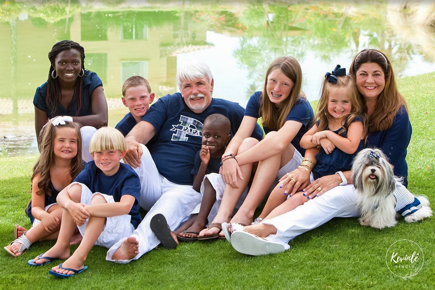 A large multigenerational family poses outdoors near a pond, smiling together with a small dog on a sunny day.