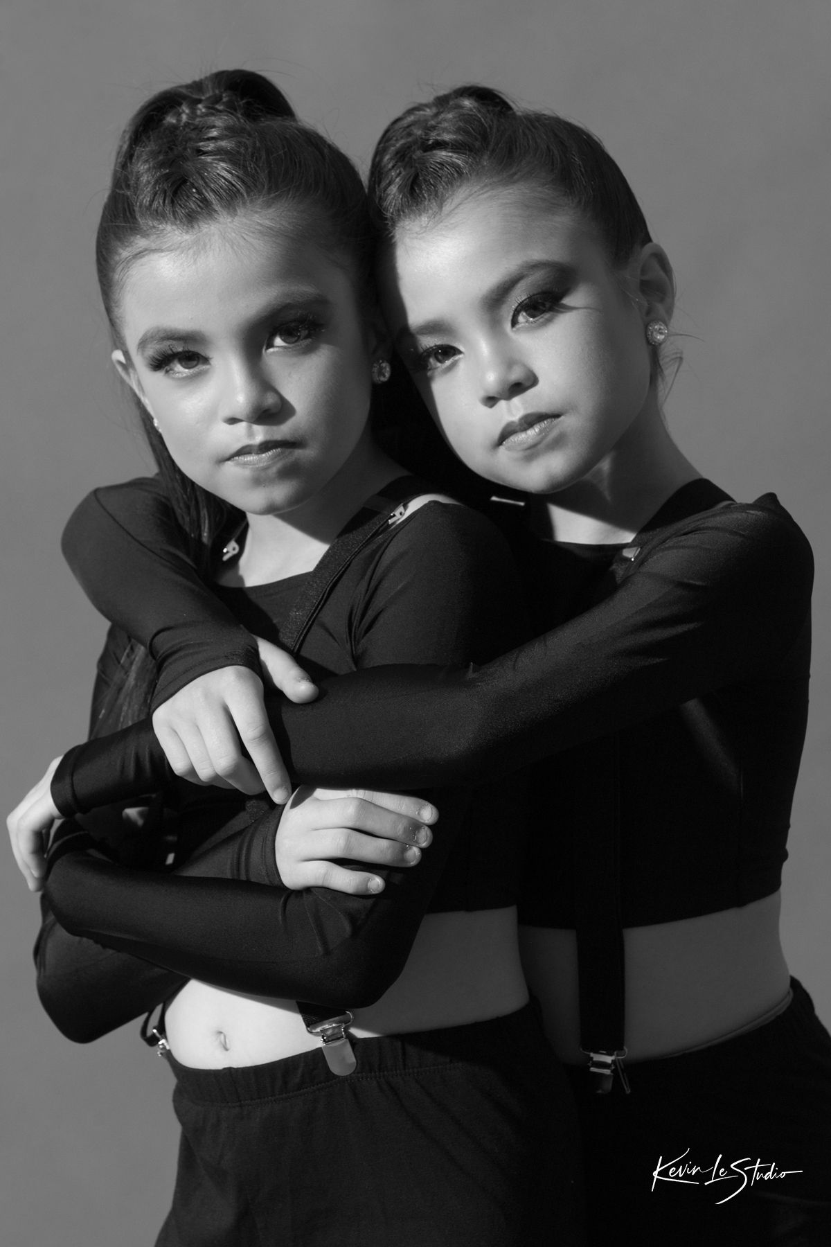 Black and white portrait of two young girls in black dance attire hugging each other in a studio setting.