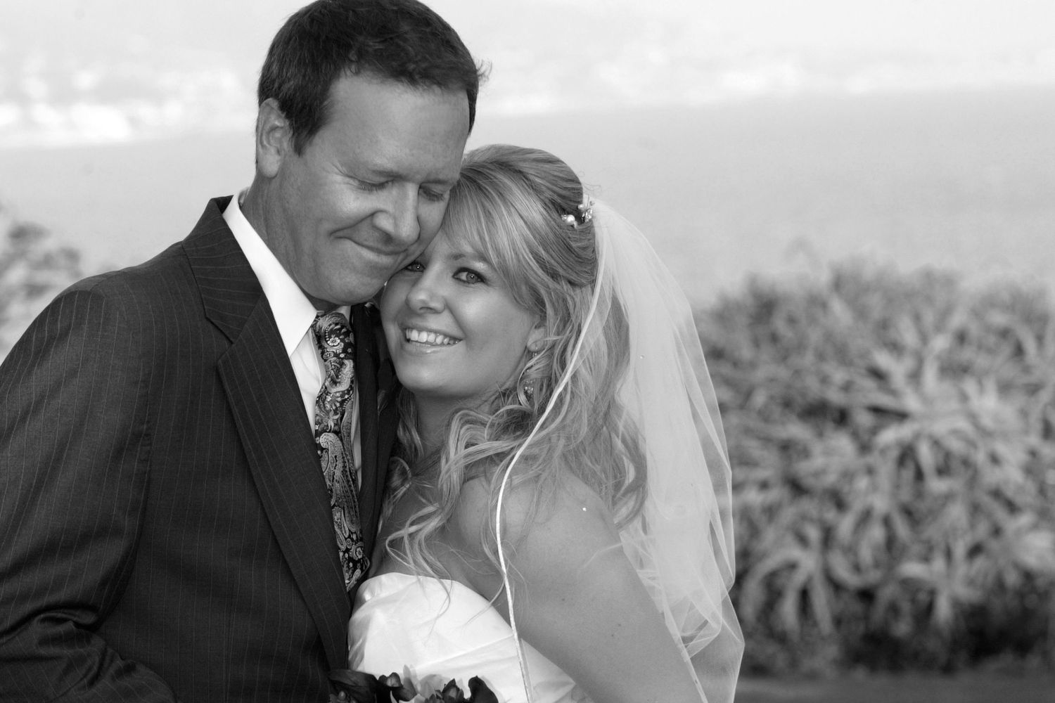 Black and white wedding photo of smiling bride in veil and groom in suit posing together outdoors.
