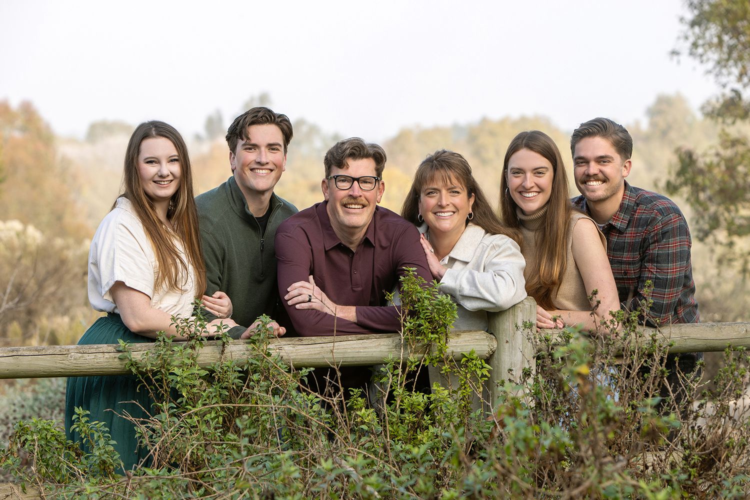 A happy family of six poses together leaning on a wooden fence surrounded by lush greenery outdoors.