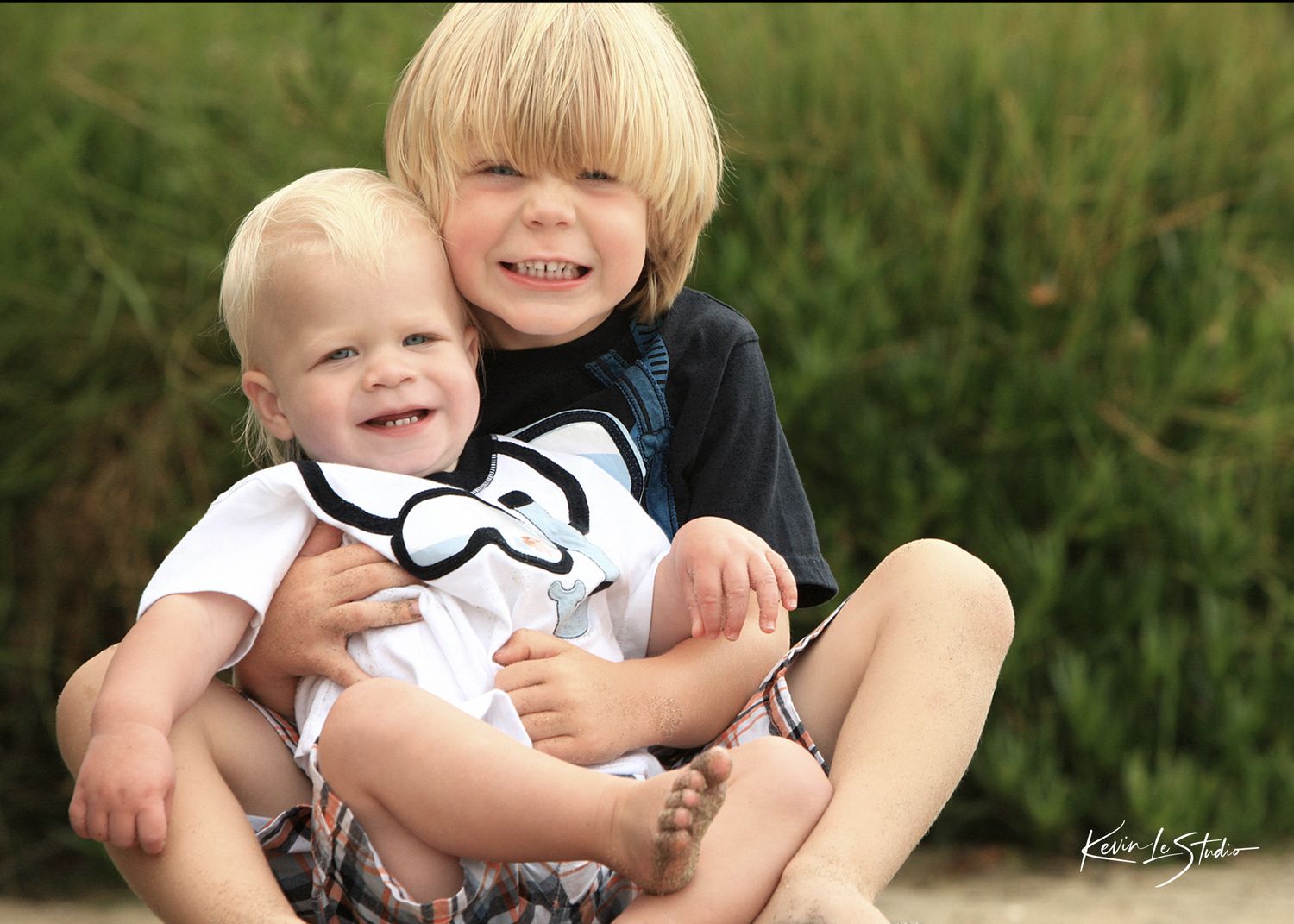 Two smiling blonde boys sitting outdoors, older brother hugging toddler holding mustache prop, with green grass background.
