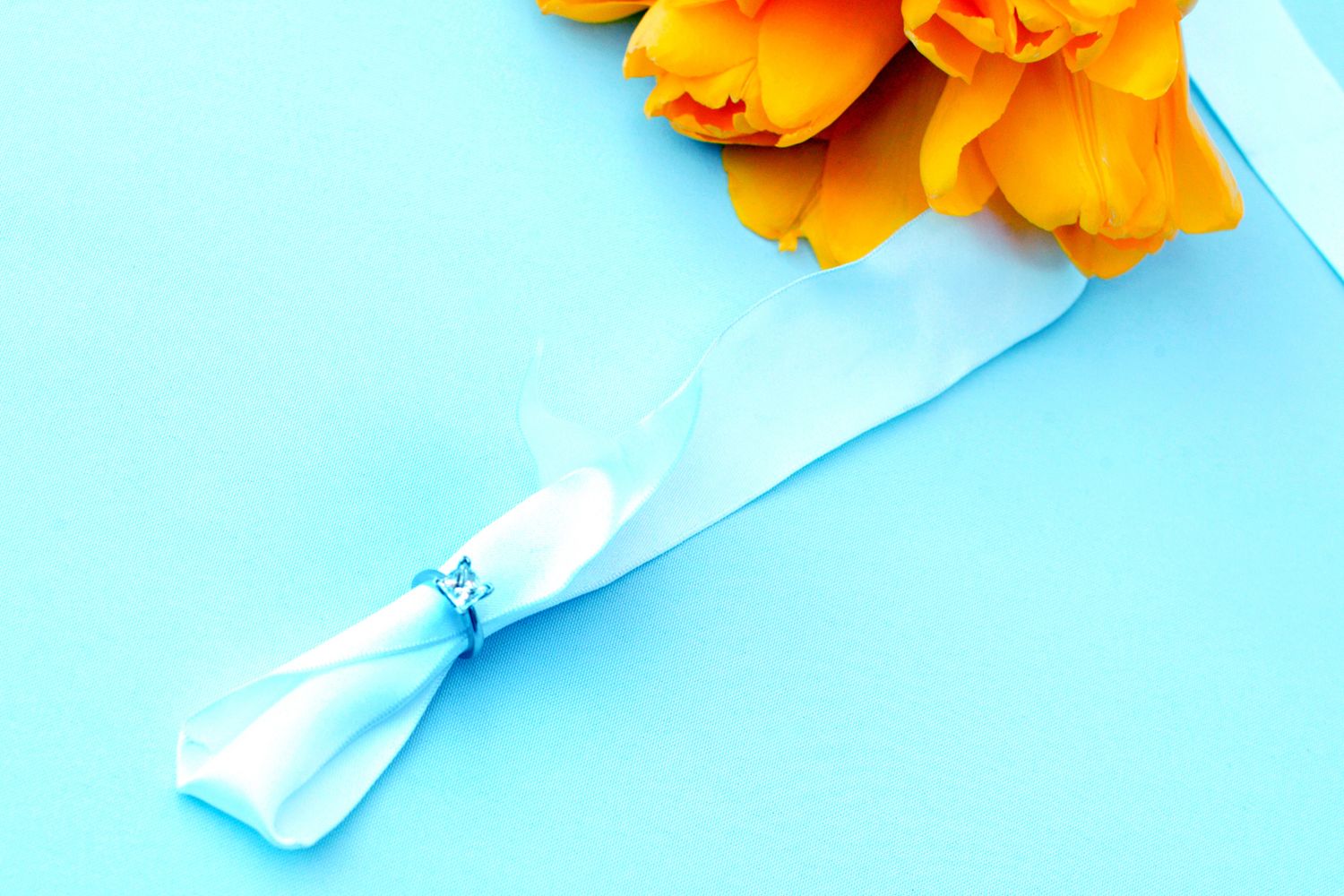 Orange tulips bouquet wrapped with white ribbon and blue diamond brooch on light blue background.