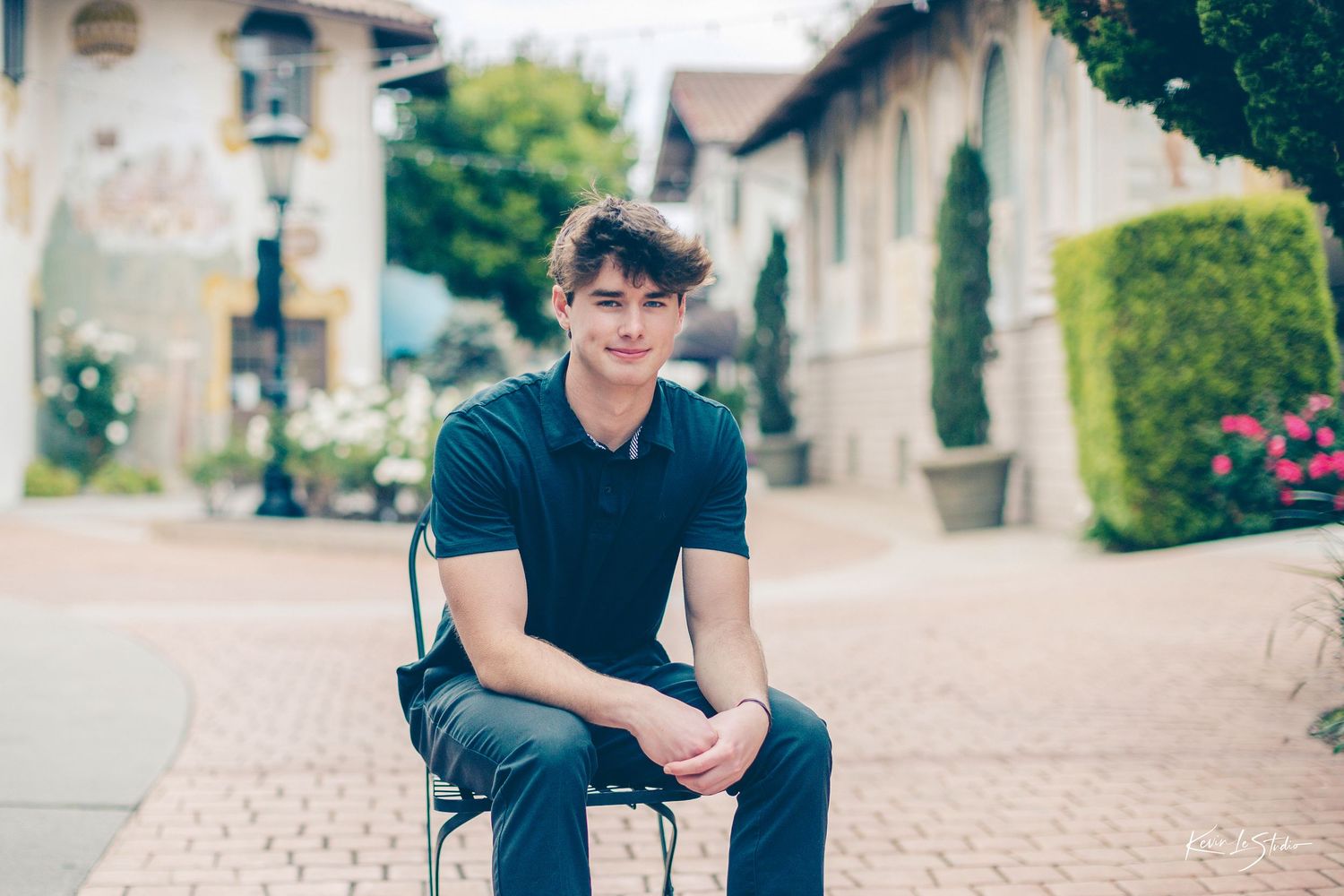Young man in dark polo shirt sitting relaxed on an iron bench in a charming outdoor village setting.