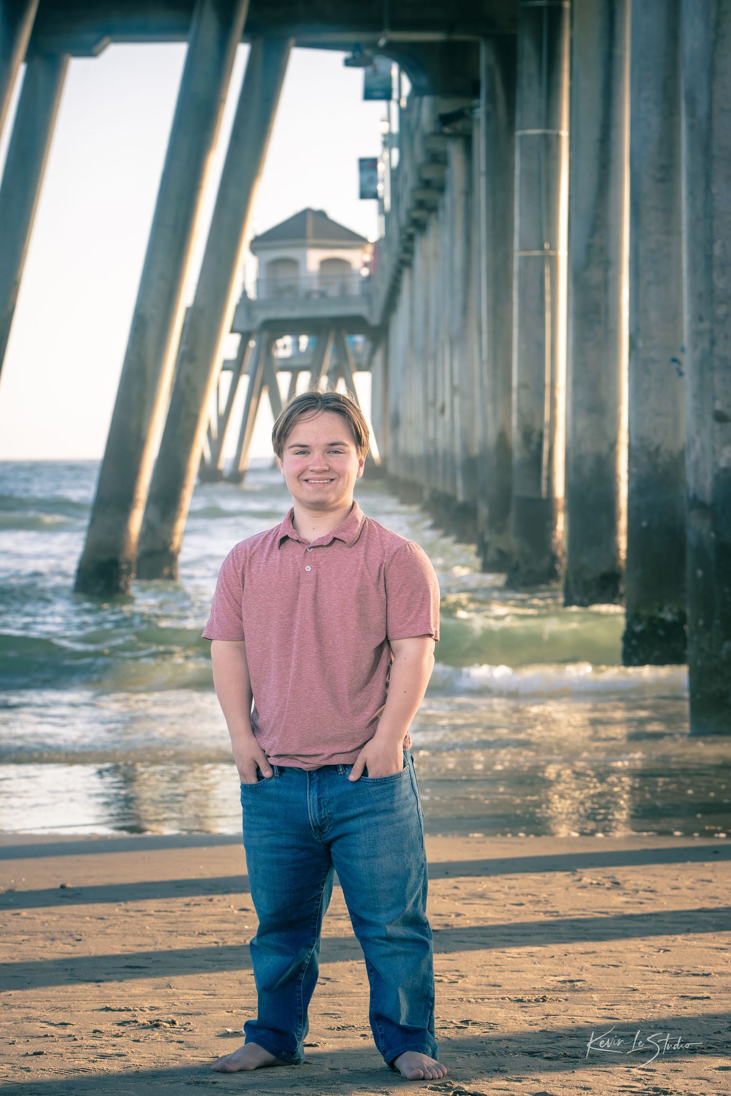 Young man smiling at beach pier in pink polo and jeans during golden hour.