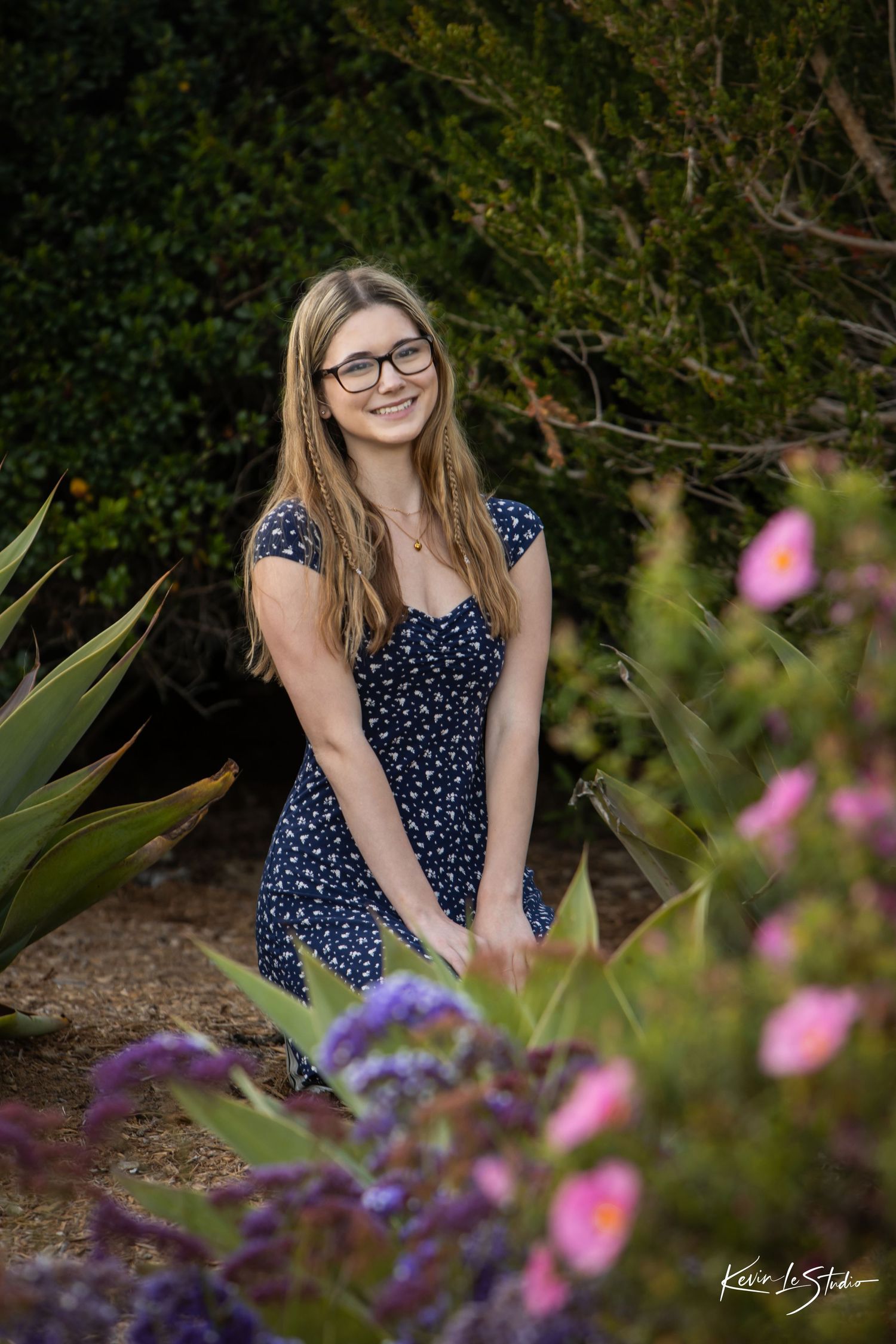 Smiling blonde woman in navy floral dress kneels among colorful garden flowers and greenery.