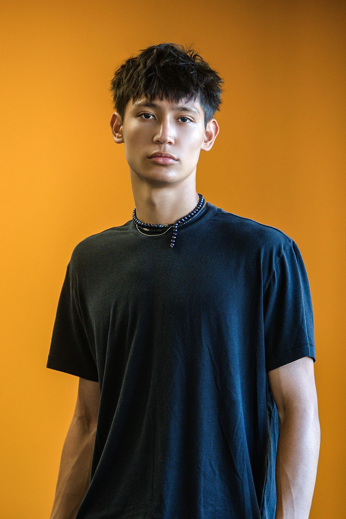 Young Asian man in dark oversized t-shirt and chain necklace poses against bright orange background.