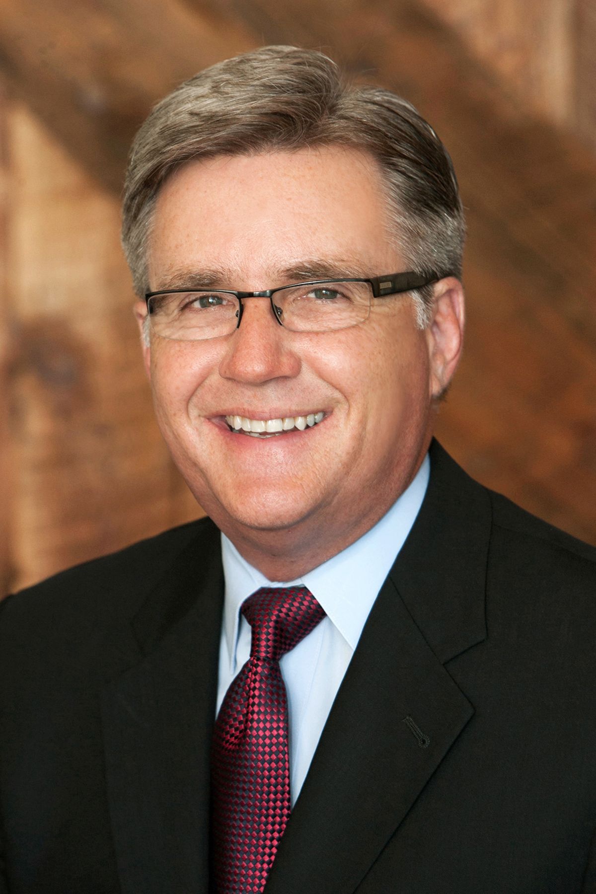 Professional headshot of a smiling older man with gray hair and glasses wearing a dark suit and red tie.