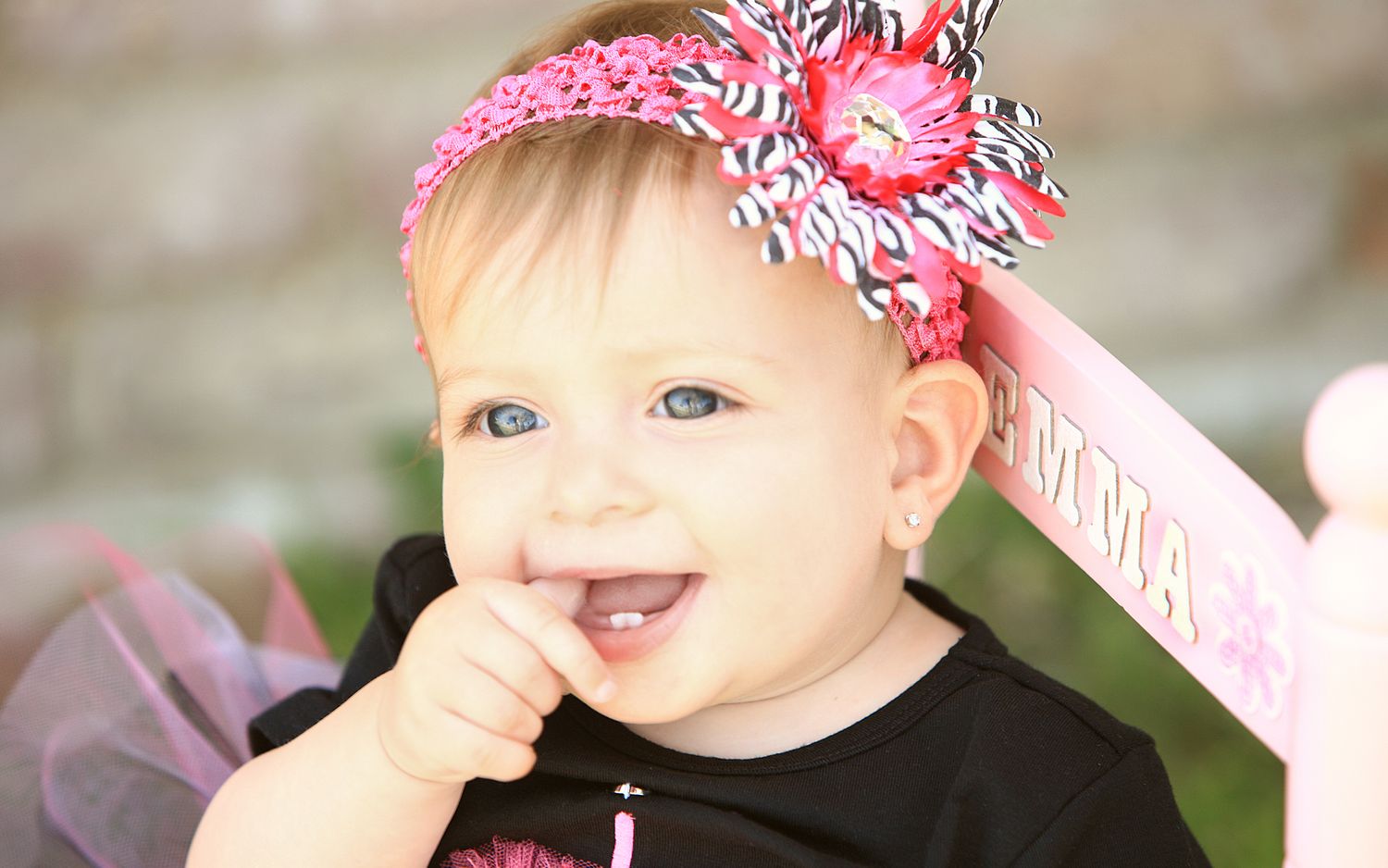 Adorable baby girl with pink floral headband smiling with finger in mouth.