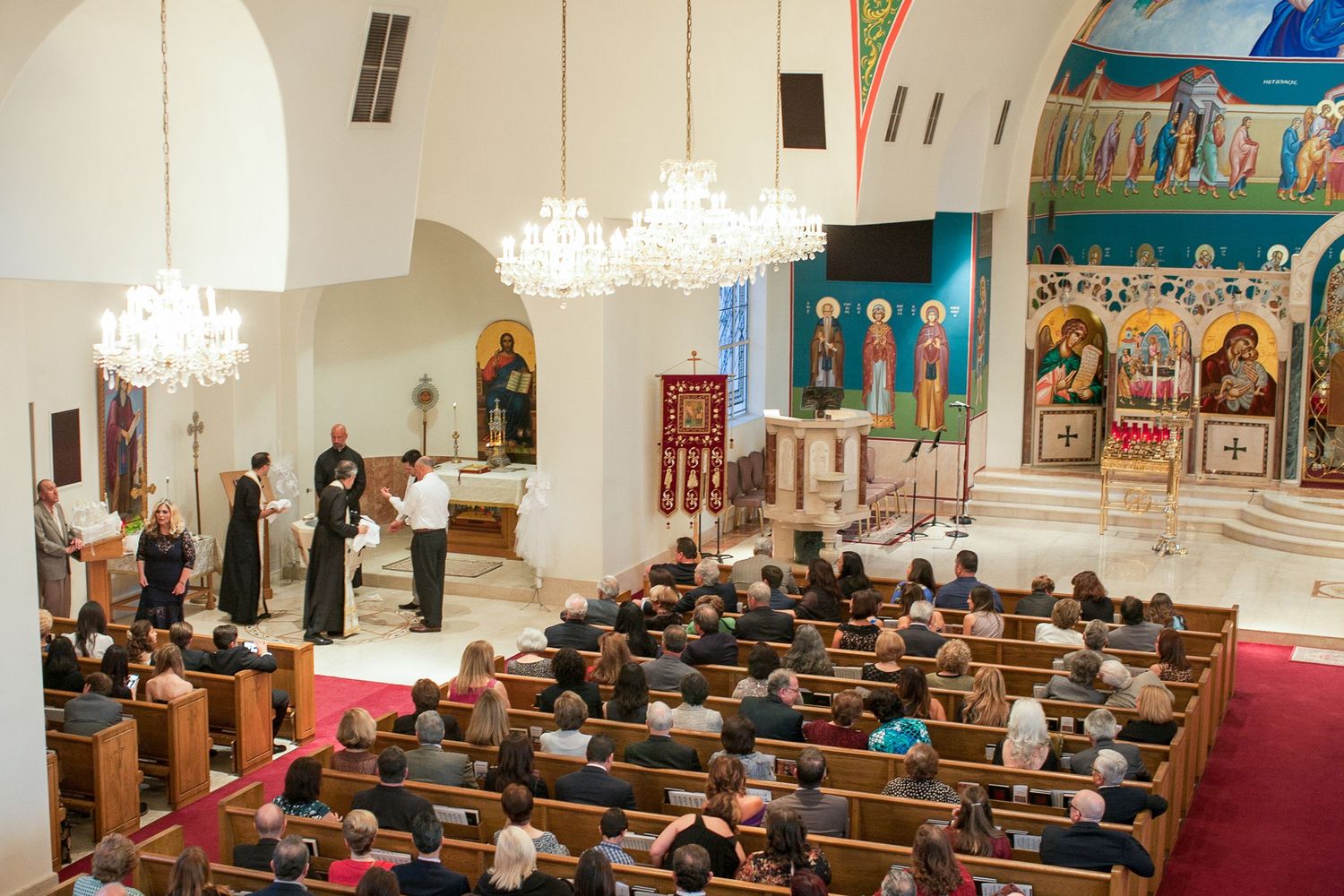 Orthodox church service with congregation seated in wooden pews, clergy at altar, chandeliers, and colorful Byzantine icons.