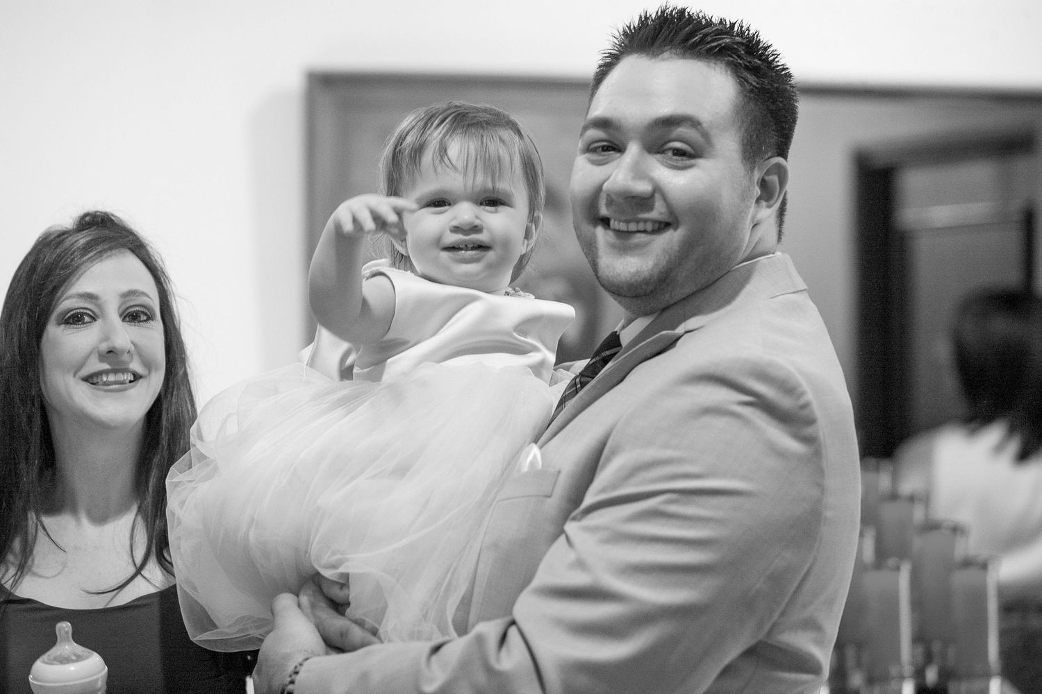 Man in gray suit smiling while holding a baby girl in white dress at a church celebration.
