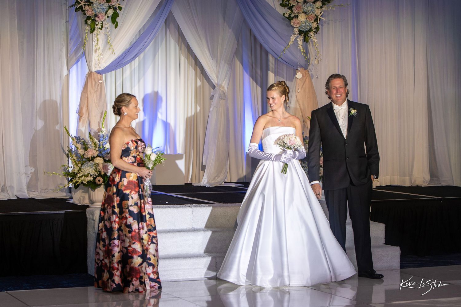 Bride in white ball gown walks down aisle with escort at elegant wedding ceremony with draped backdrop.