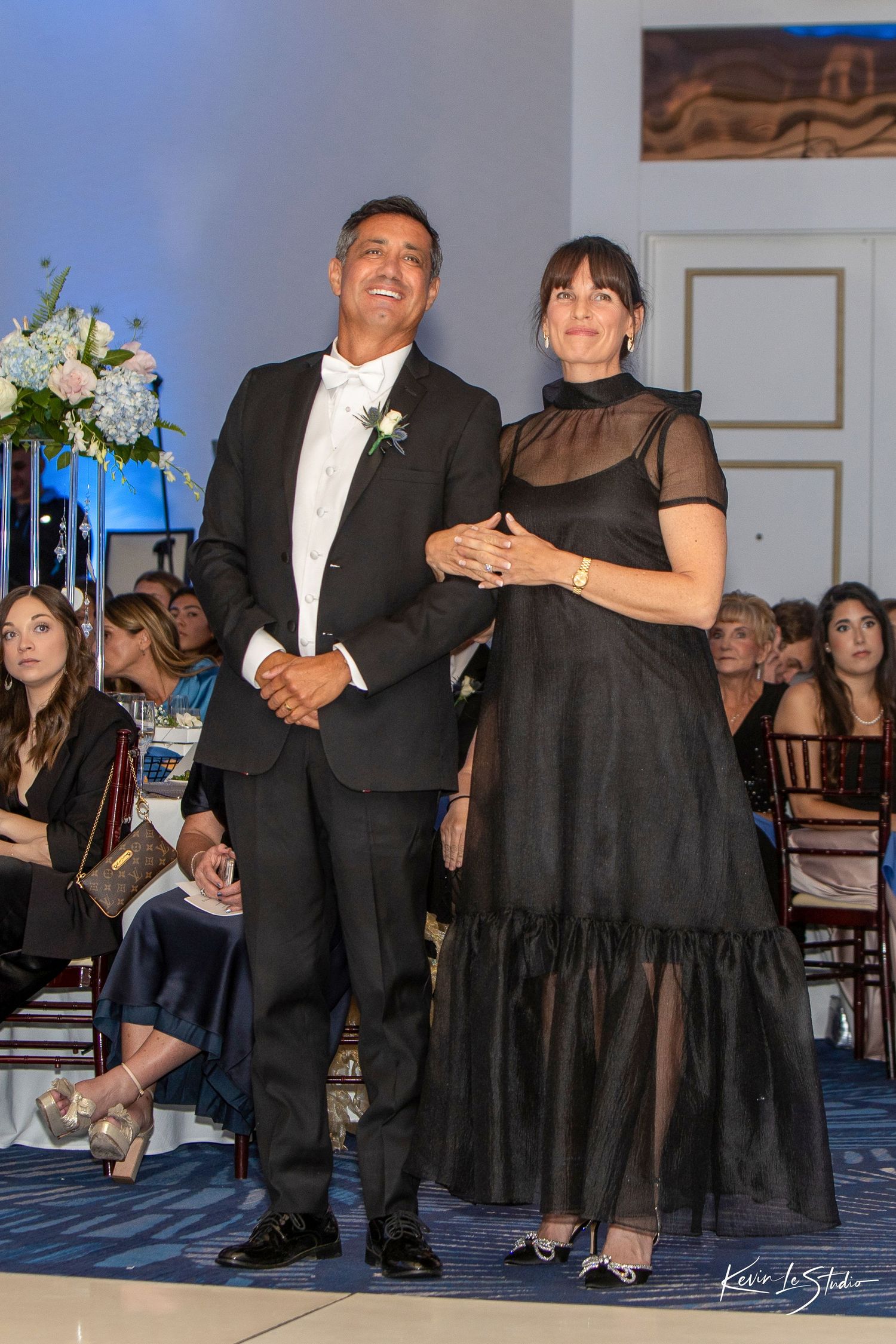 Elegant couple in black formal attire smiling at a wedding reception, man in tuxedo with boutonniere, woman in sheer gown.