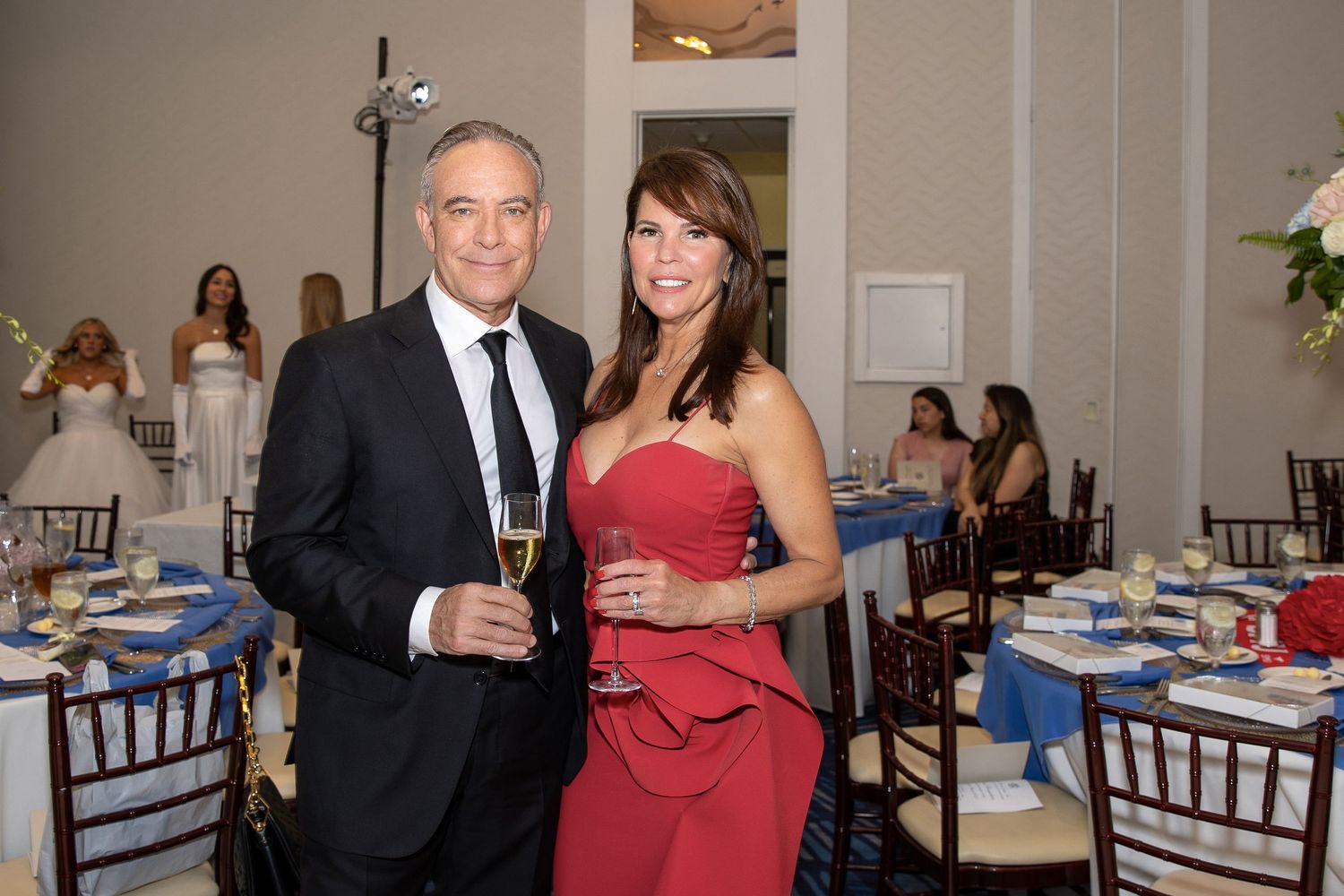 Elegant couple in formal attire holding champagne glasses at a gala dinner event with decorated tables.