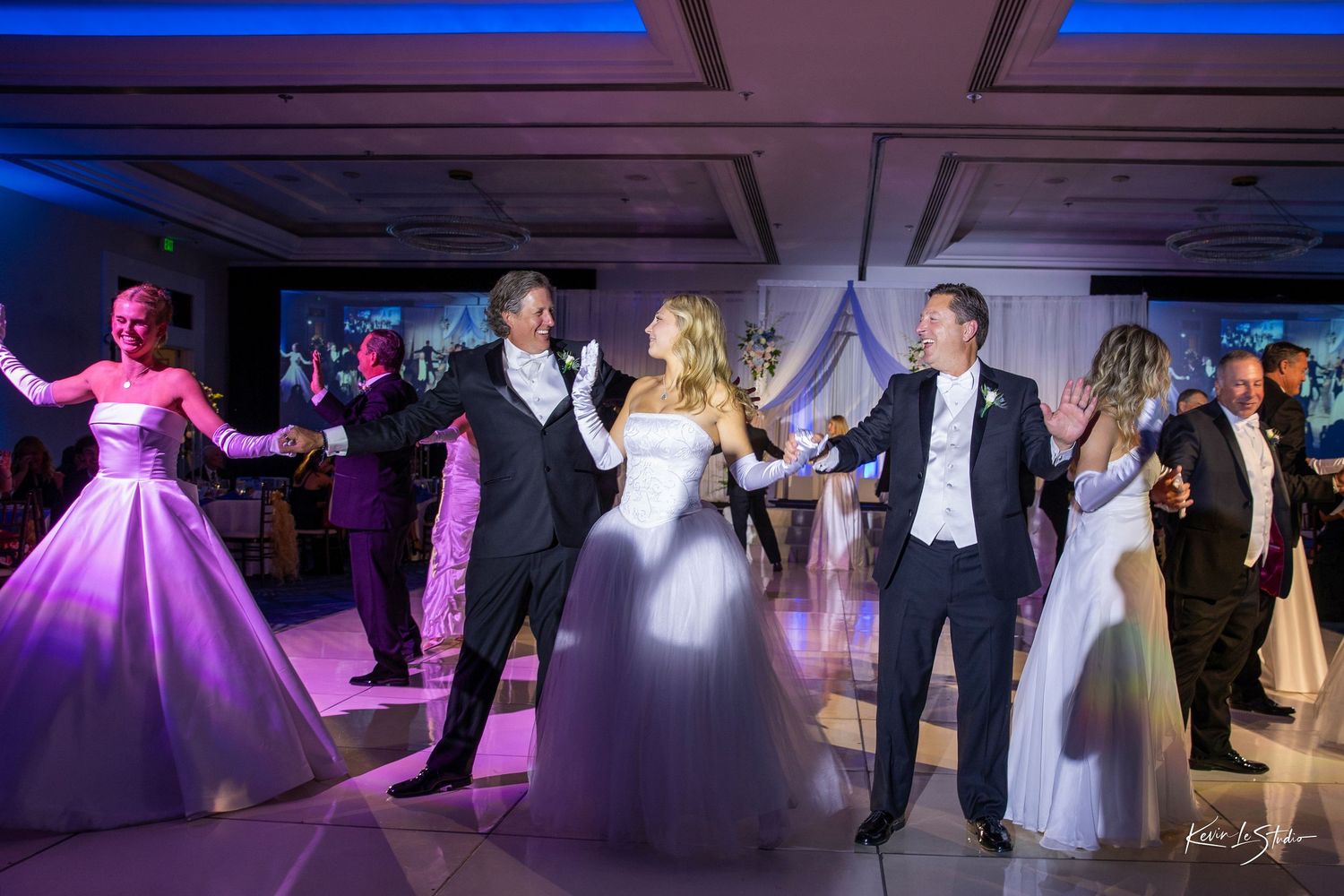 Wedding reception dance floor with bride in white gown, groom in tuxedo, and guests dancing under purple lights.