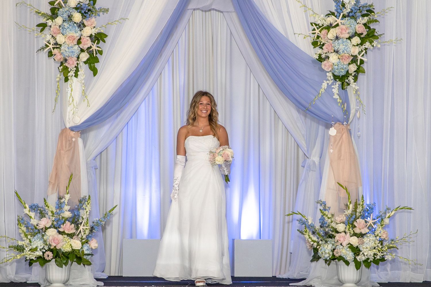 Bride in white strapless gown holding bouquet stands under blue draped arch with pink and blue floral arrangements.
