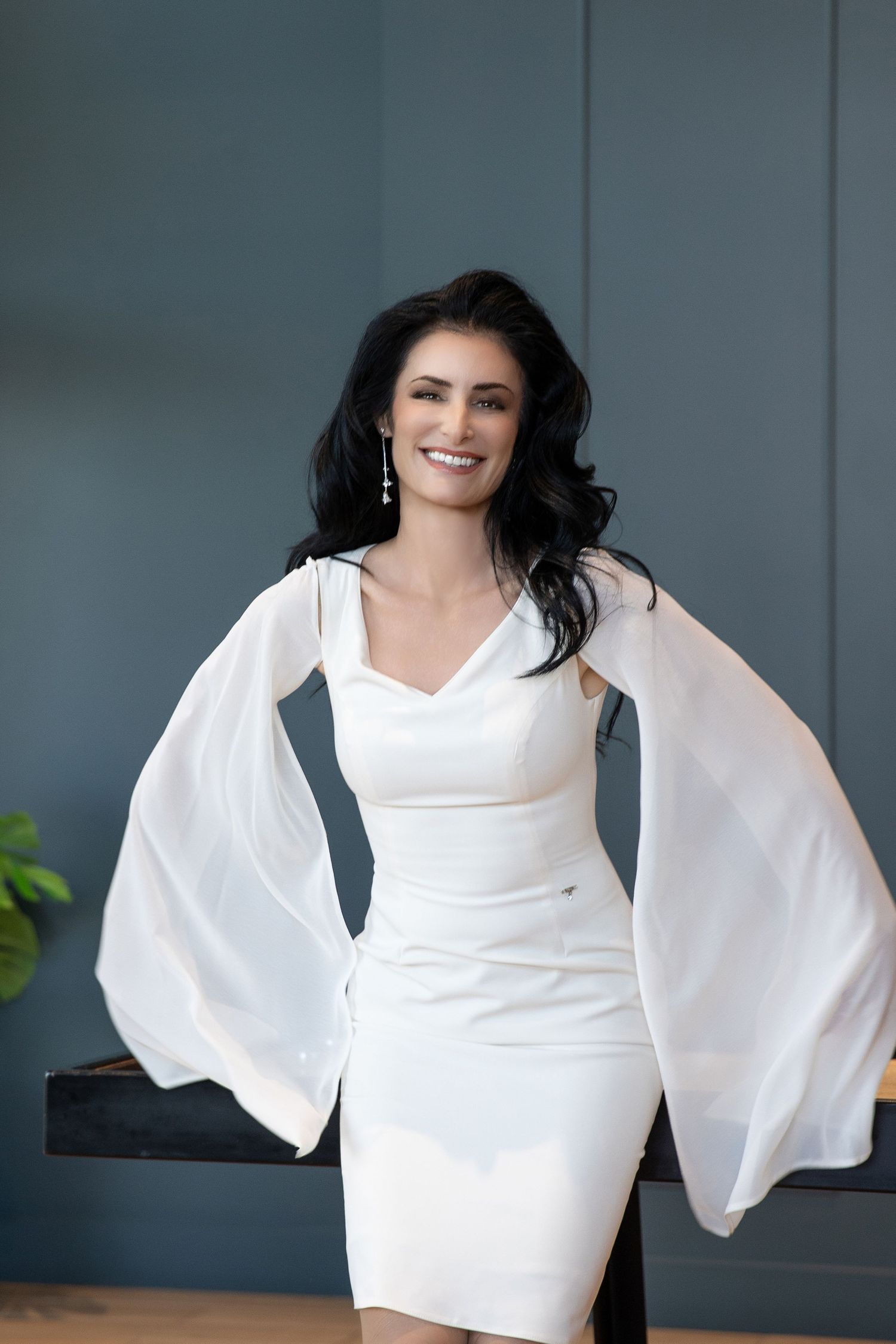 Smiling brunette woman in elegant white dress with flowing sheer sleeves posing confidently indoors.