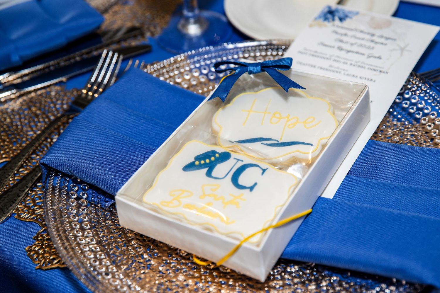 UC graduation decorated sugar cookies in blue and gold displayed in a gift box on an elegant sequined table setting.