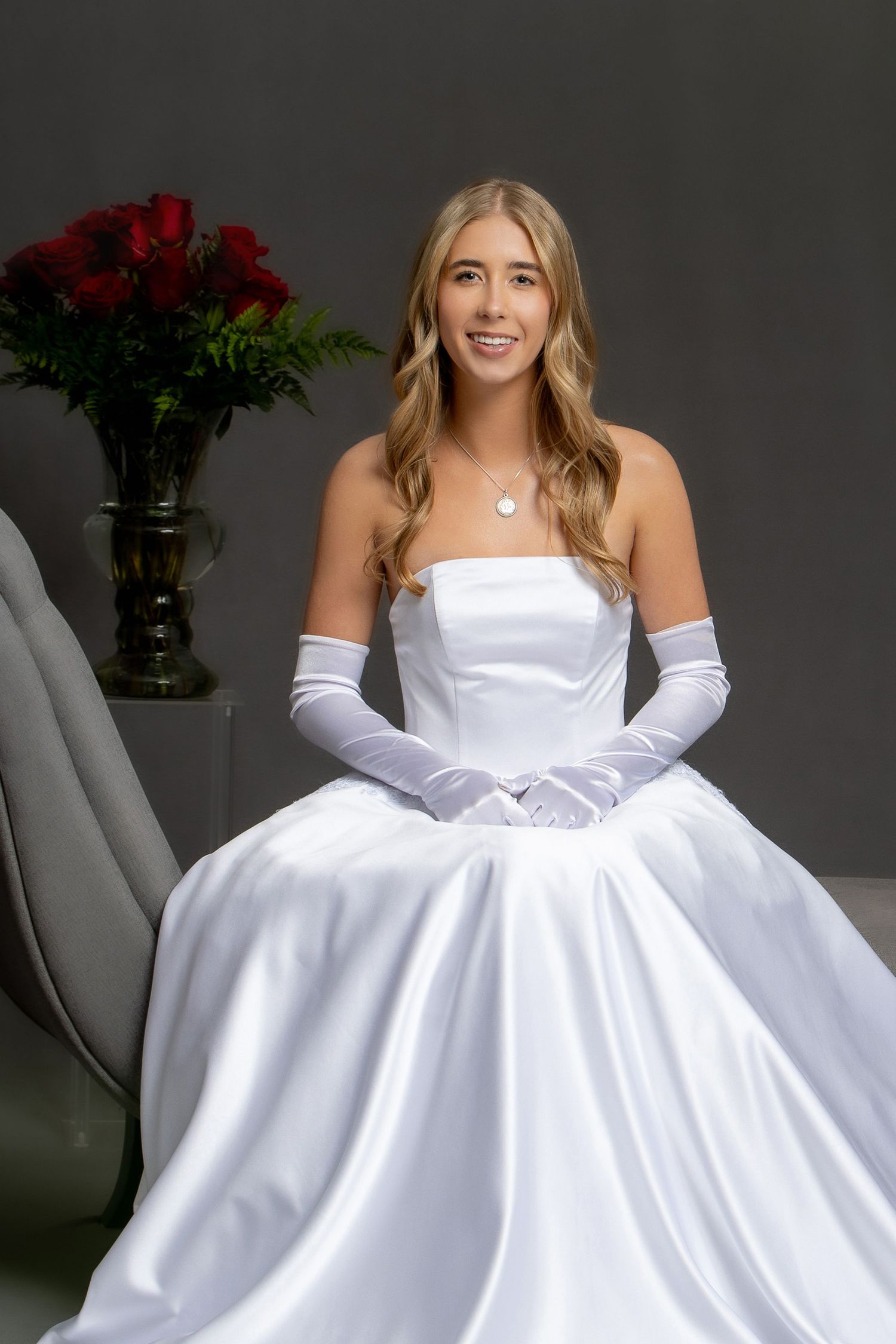 Young woman in white strapless satin ball gown with white gloves seated near red roses bouquet.