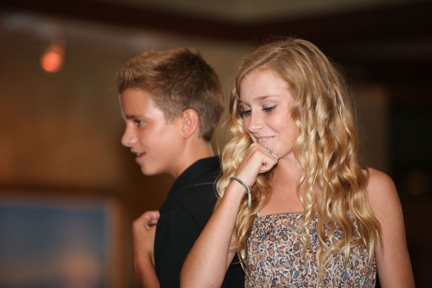 Two teenagers, a boy and a blonde girl, stand together at an indoor event, both looking downward thoughtfully.