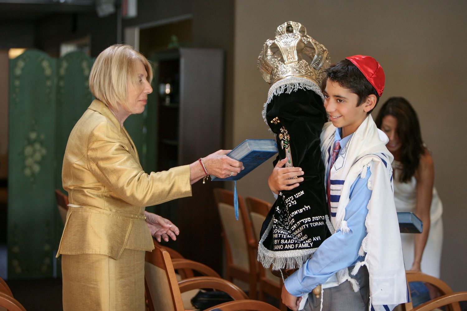 Boy in tallit holds Torah scroll during Bar Mitzvah ceremony while woman presents him with a gift.