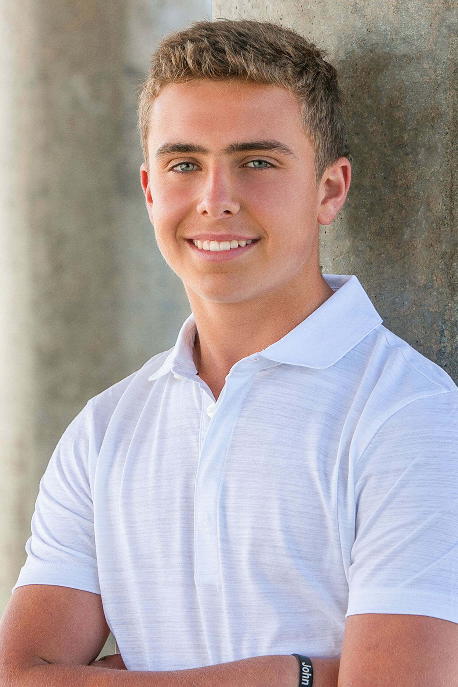 Smiling young man with blue eyes wearing a white polo shirt, arms crossed, posing outdoors against a concrete wall.