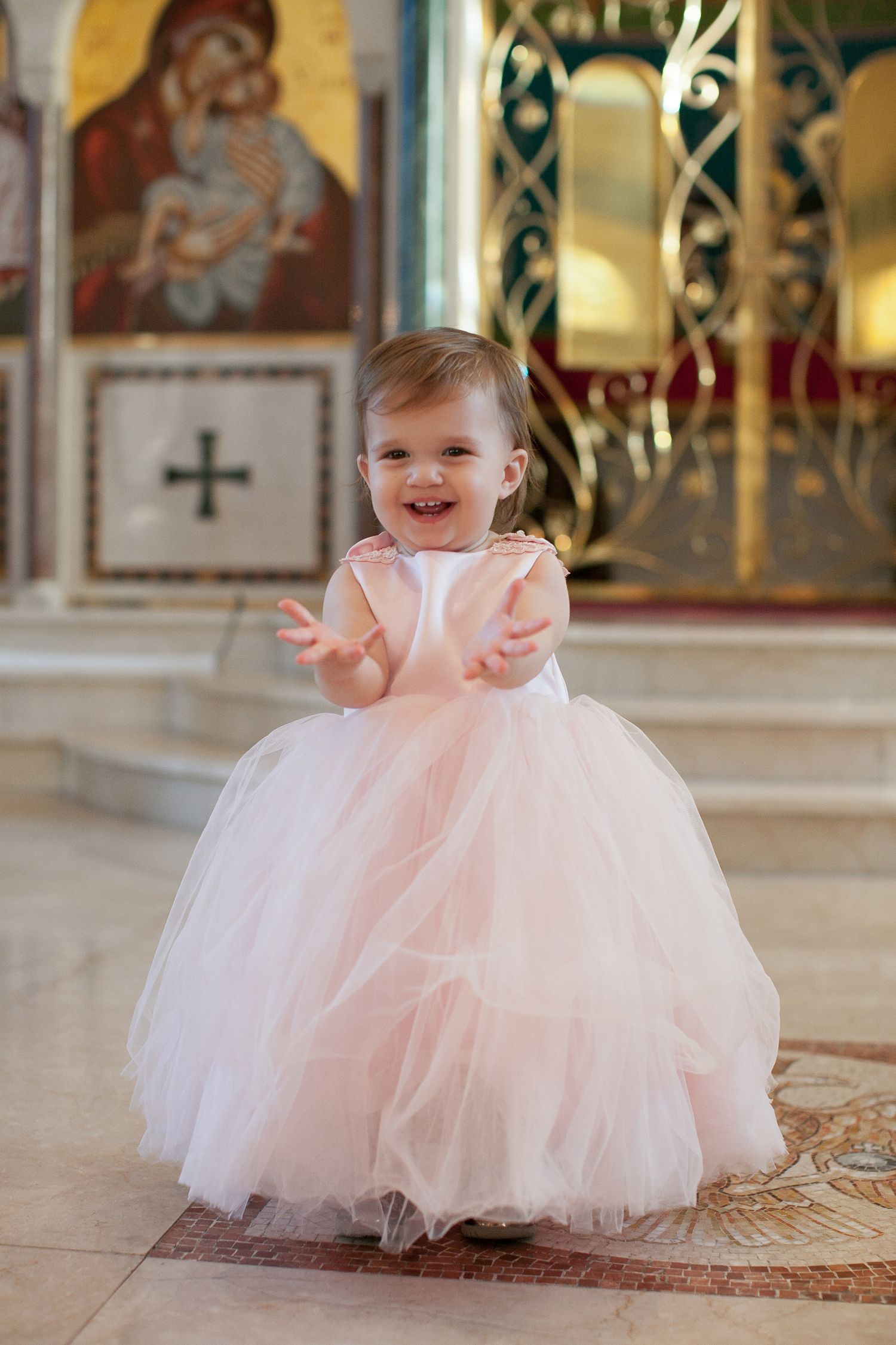 Smiling toddler girl in pink tulle dress clapping hands inside an Orthodox church.