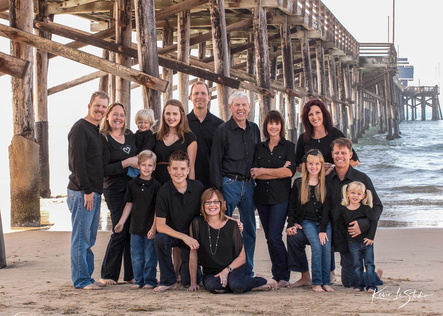 Large family portrait on sandy beach near wooden pier, all wearing black tops and jeans, smiling together.