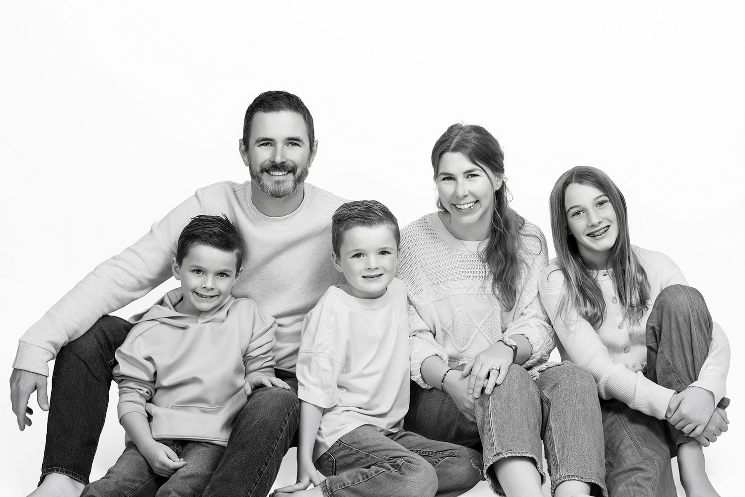 Black and white family portrait of two adults and three children sitting together, smiling warmly in a studio setting.