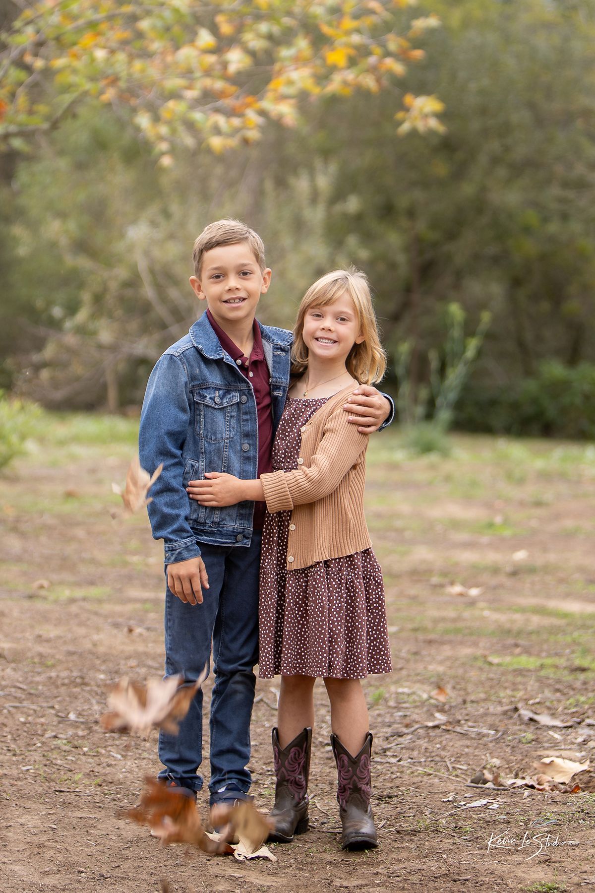 Two children pose outdoors in fall, boy in denim jacket hugging smiling girl in brown dress and cowboy boots.