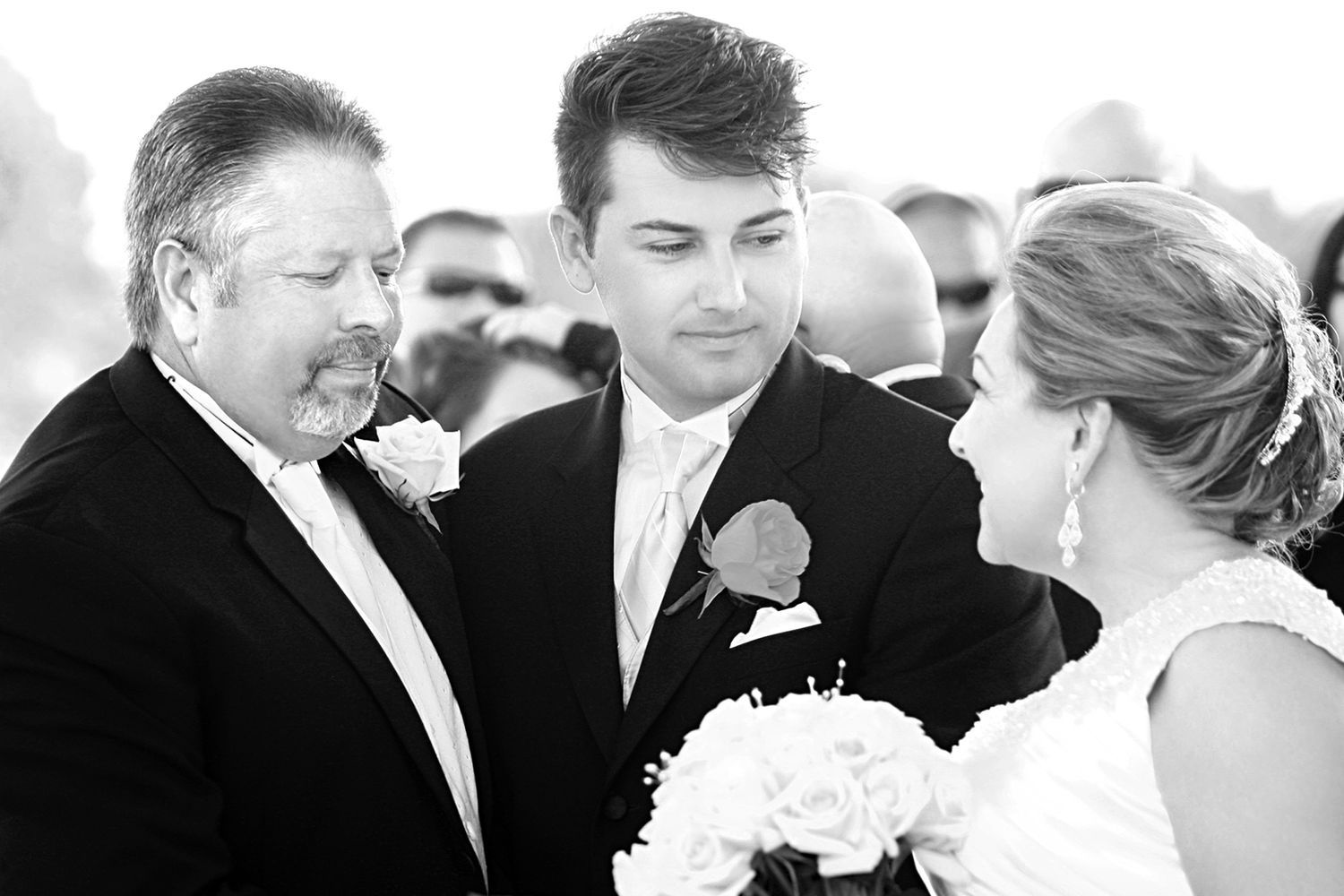 Groom and older man share an emotional moment with the bride during a black and white wedding ceremony.