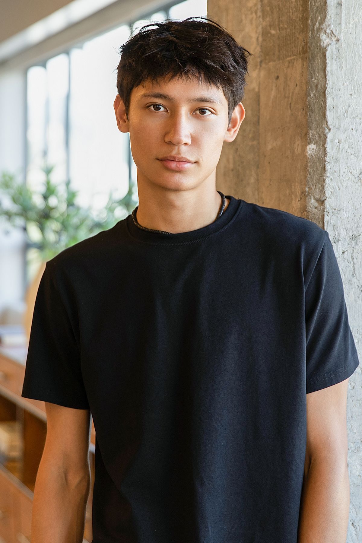 Young Asian man with short dark hair wearing a black t-shirt, standing against a concrete column indoors.