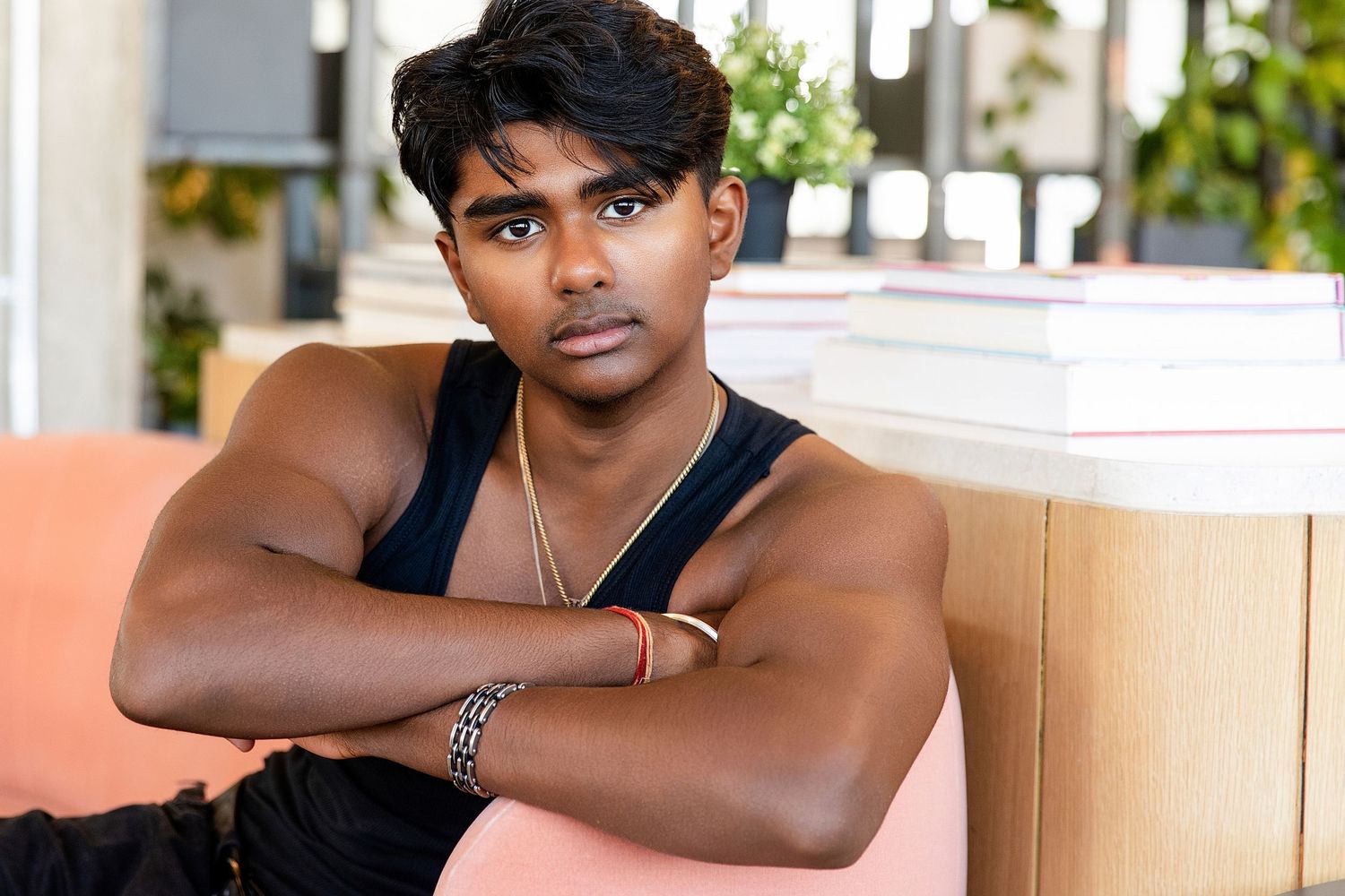 Young muscular man with crossed arms wearing black tank top and gold chain, sitting in modern interior.
