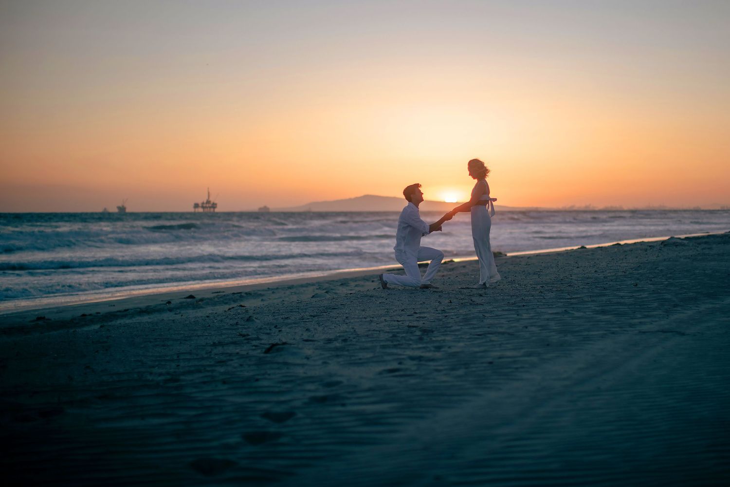 Man proposing to woman on beach at golden sunset with ocean backdrop.
