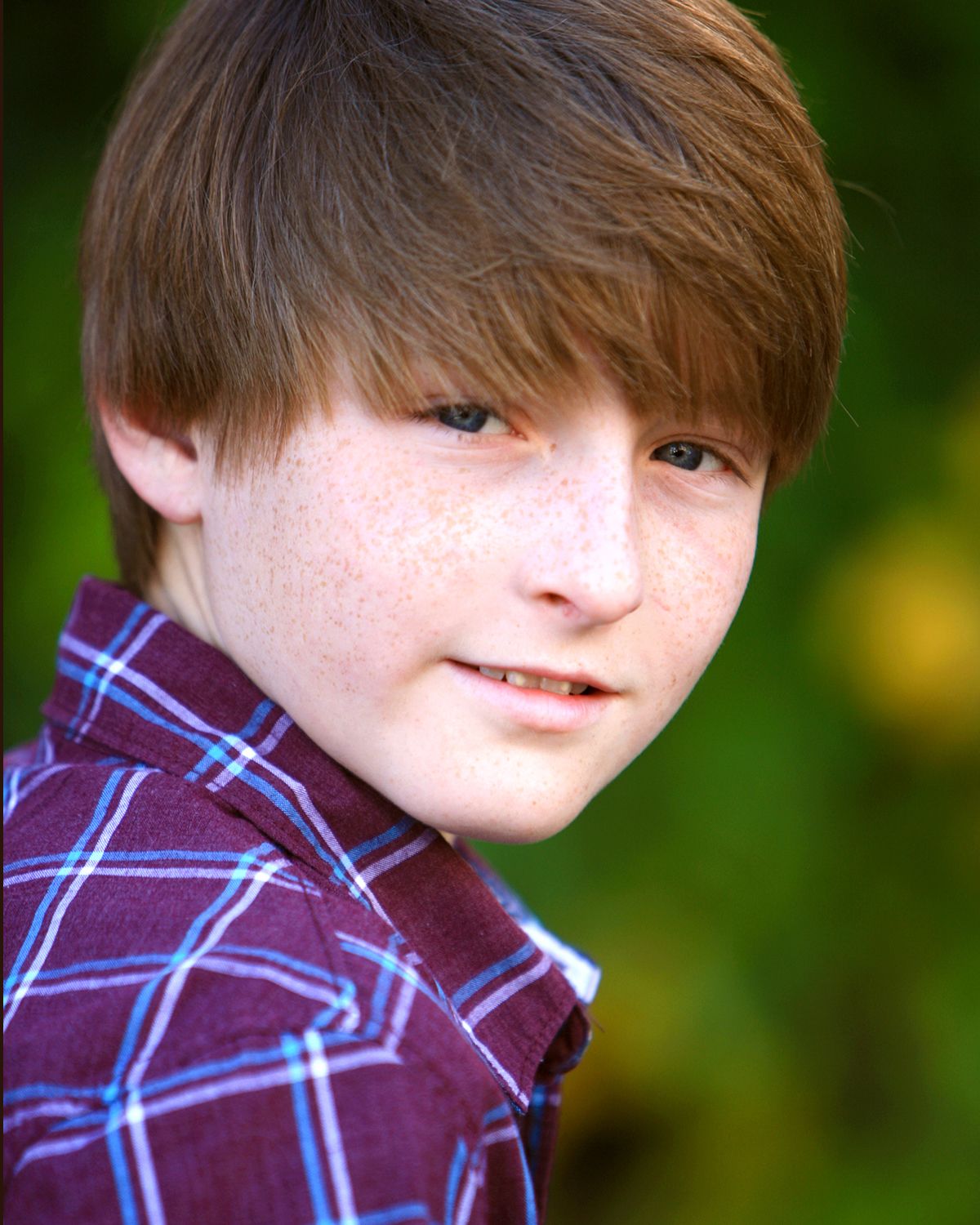 Freckled red-haired boy wearing a plaid shirt smiling in an outdoor professional headshot photo.