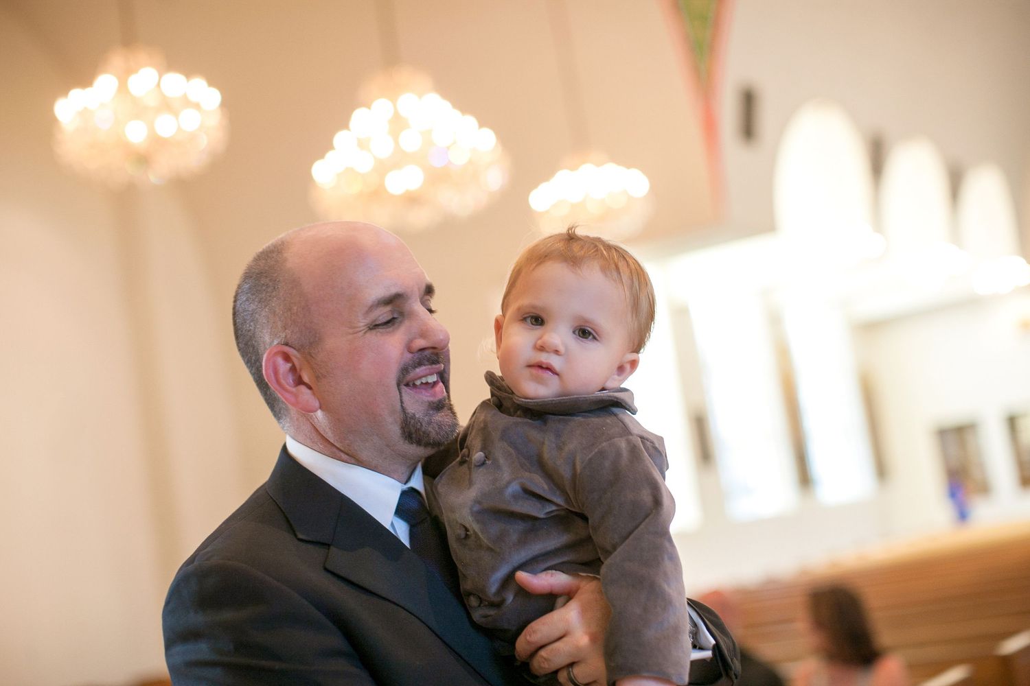 Man in suit holding a baby in a brown jacket inside an elegant church with chandeliers.