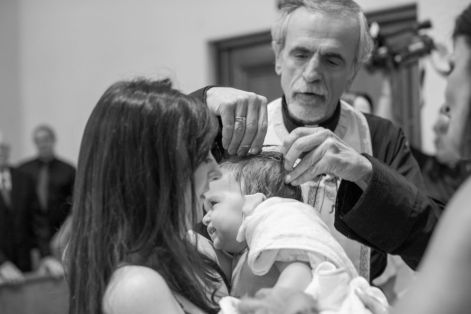 Priest performing a baptism ceremony, pouring holy water on an infant held by a woman in a church.