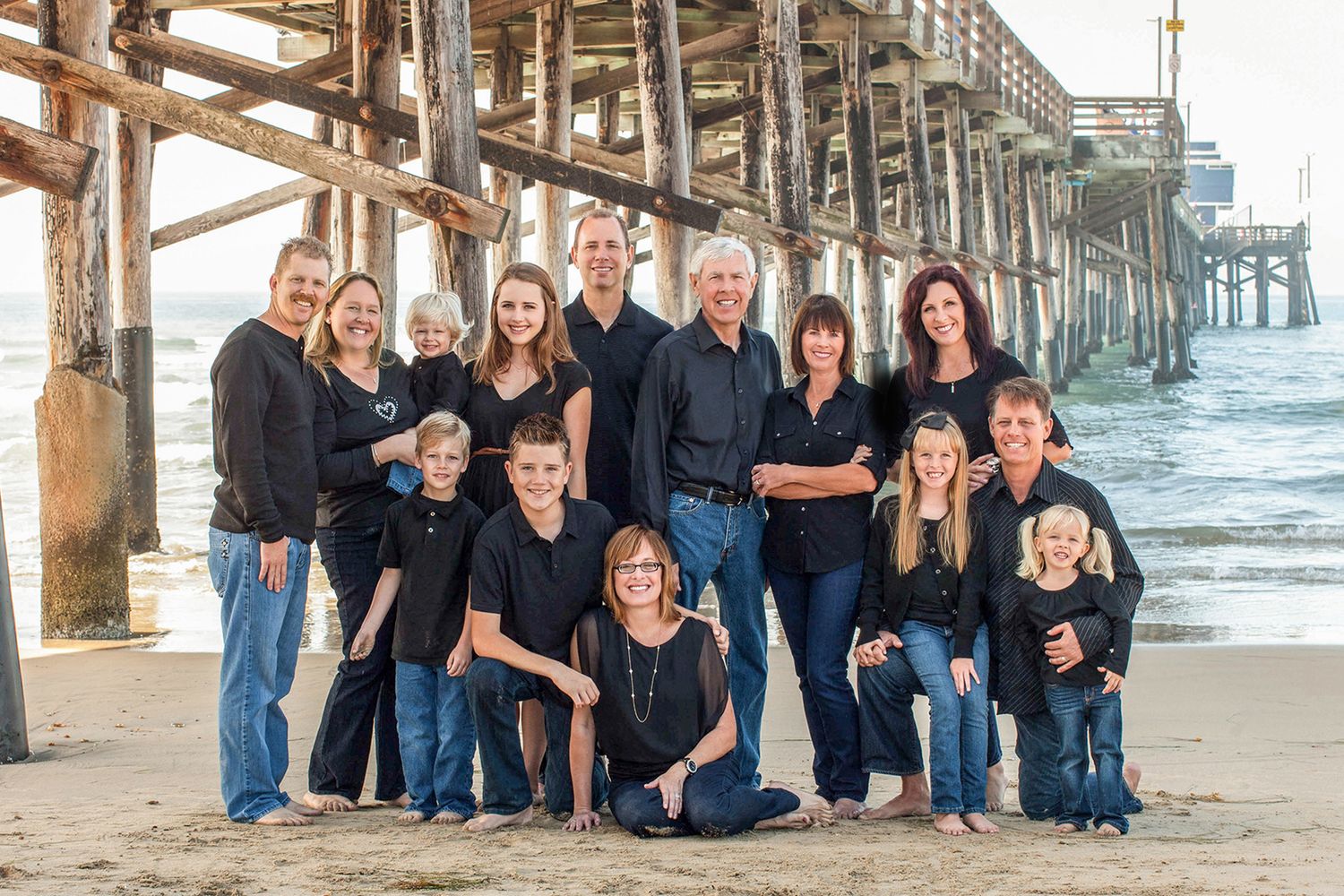 Large family group portrait on sandy beach near wooden pier, all wearing coordinating navy blue outfits.