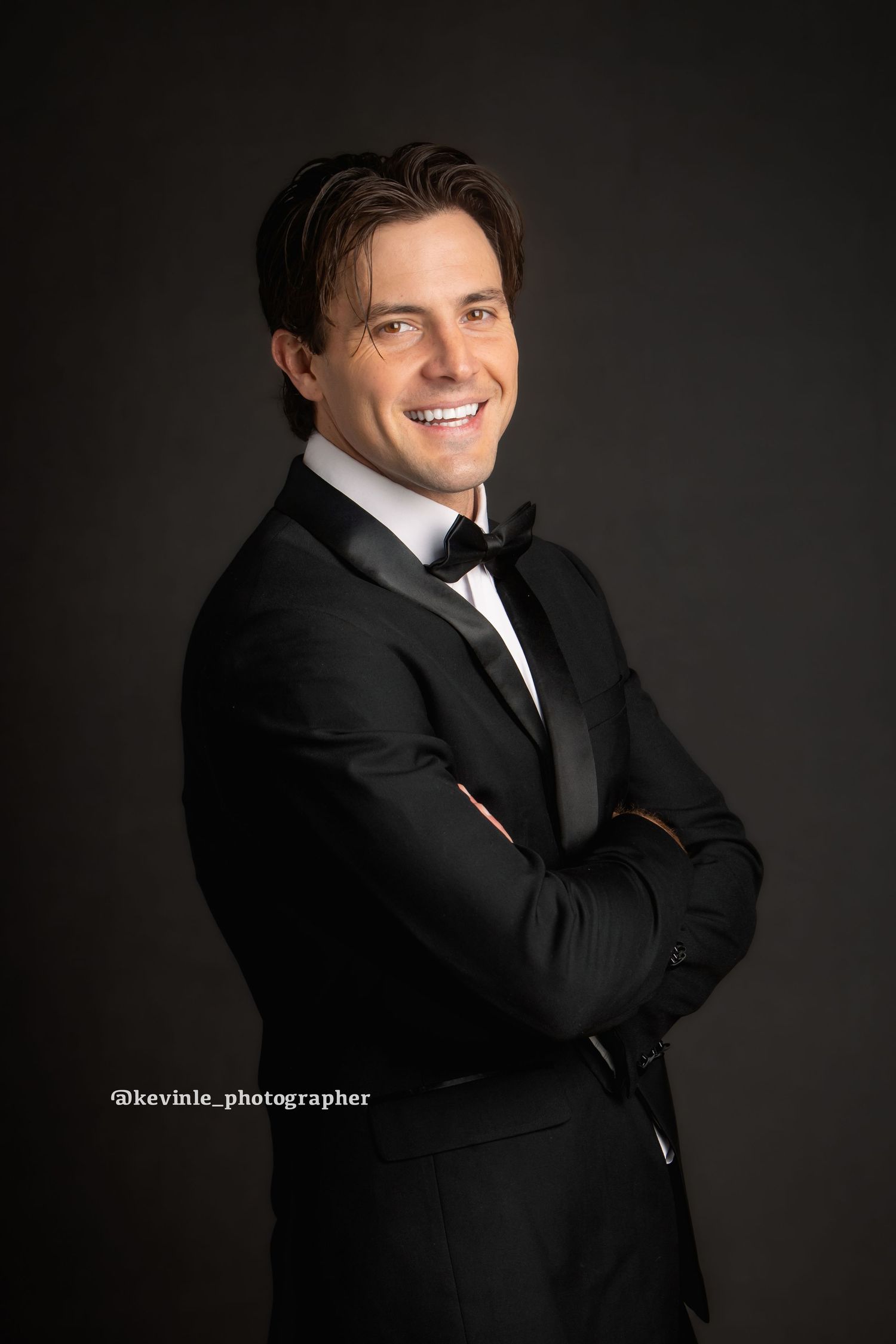 Man in black tuxedo with bow tie smiling confidently with arms crossed against dark background.