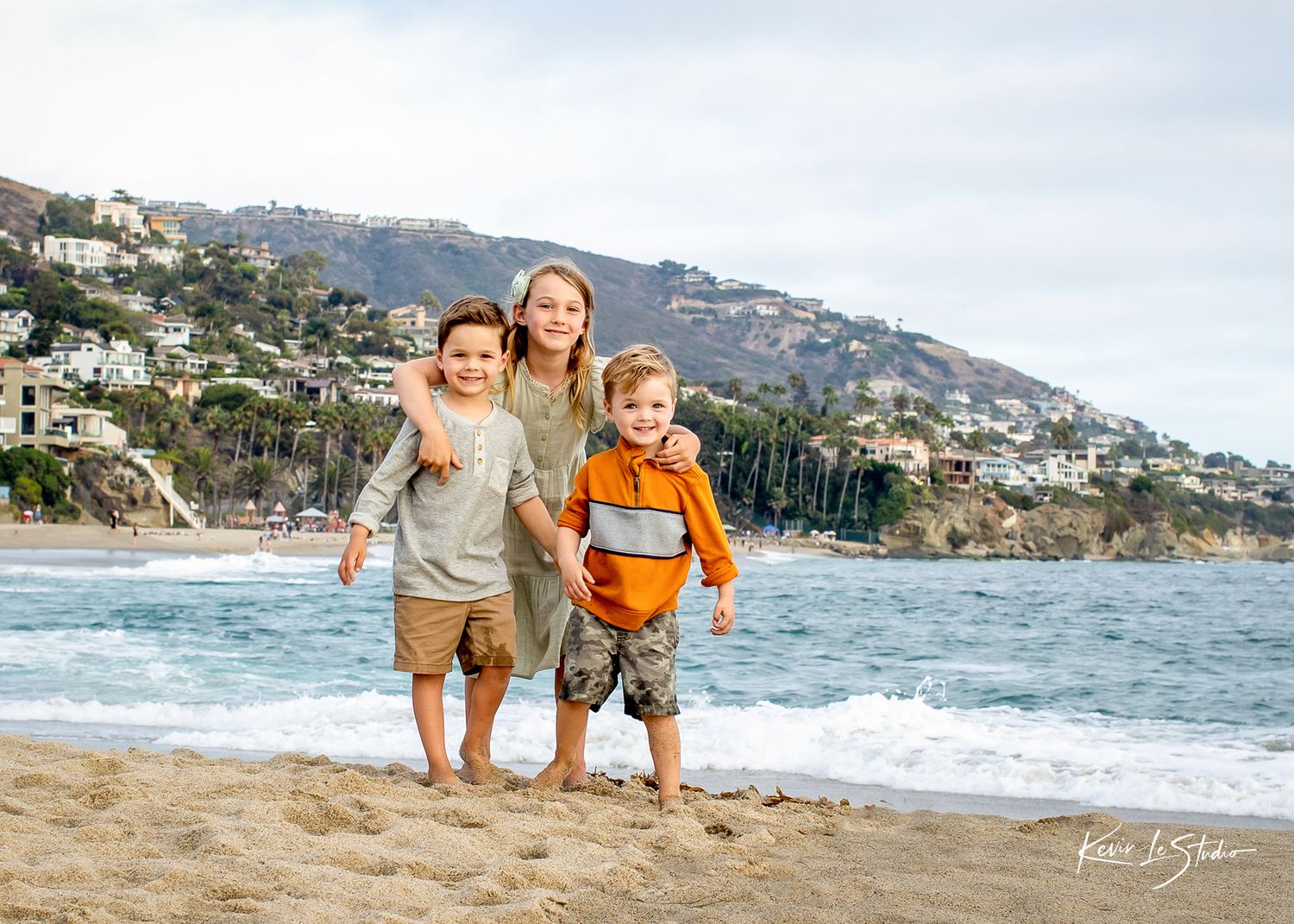 Three happy children posing together on a sandy beach with ocean waves and hillside homes behind them.
