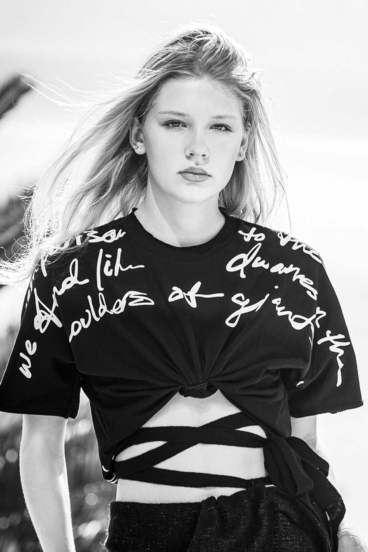 Black and white portrait of a young blonde woman in a black script-print knotted crop top, hair blowing in the wind.